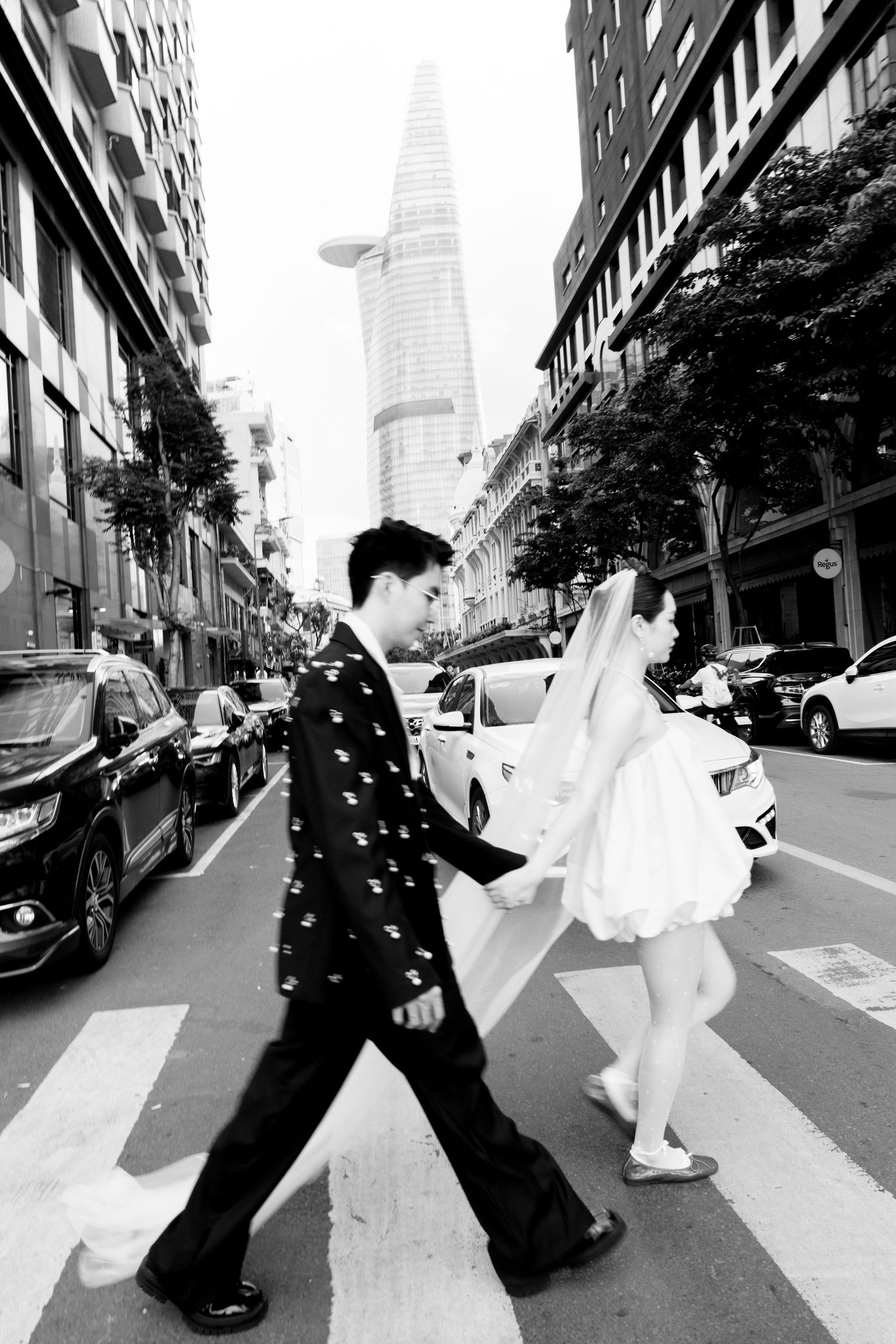 Black and white photo of couple crossing a city street holding hands with skyscraper in background.