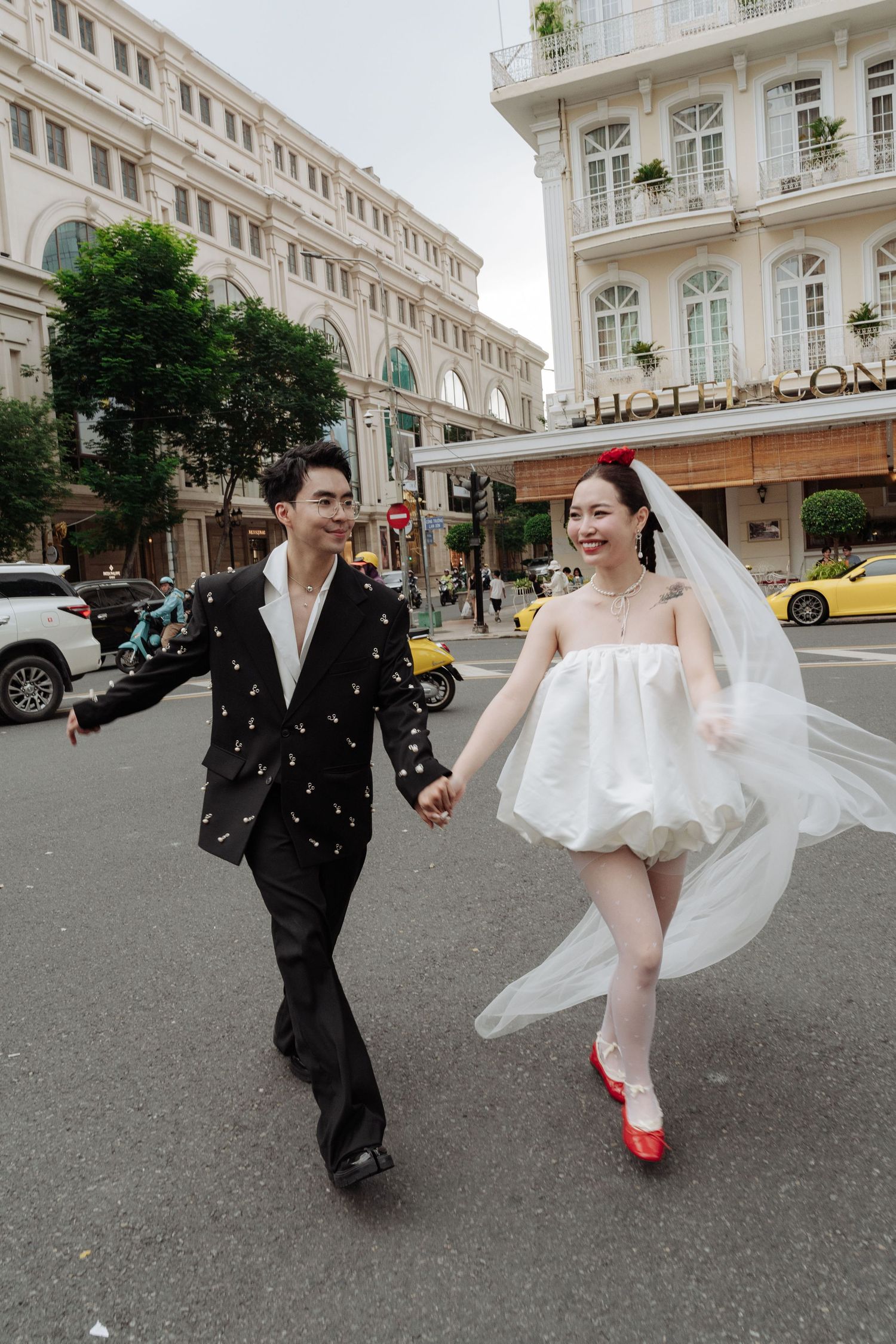 Couple in wedding attire running across a city street, bride in white mini dress with veil and red shoes.