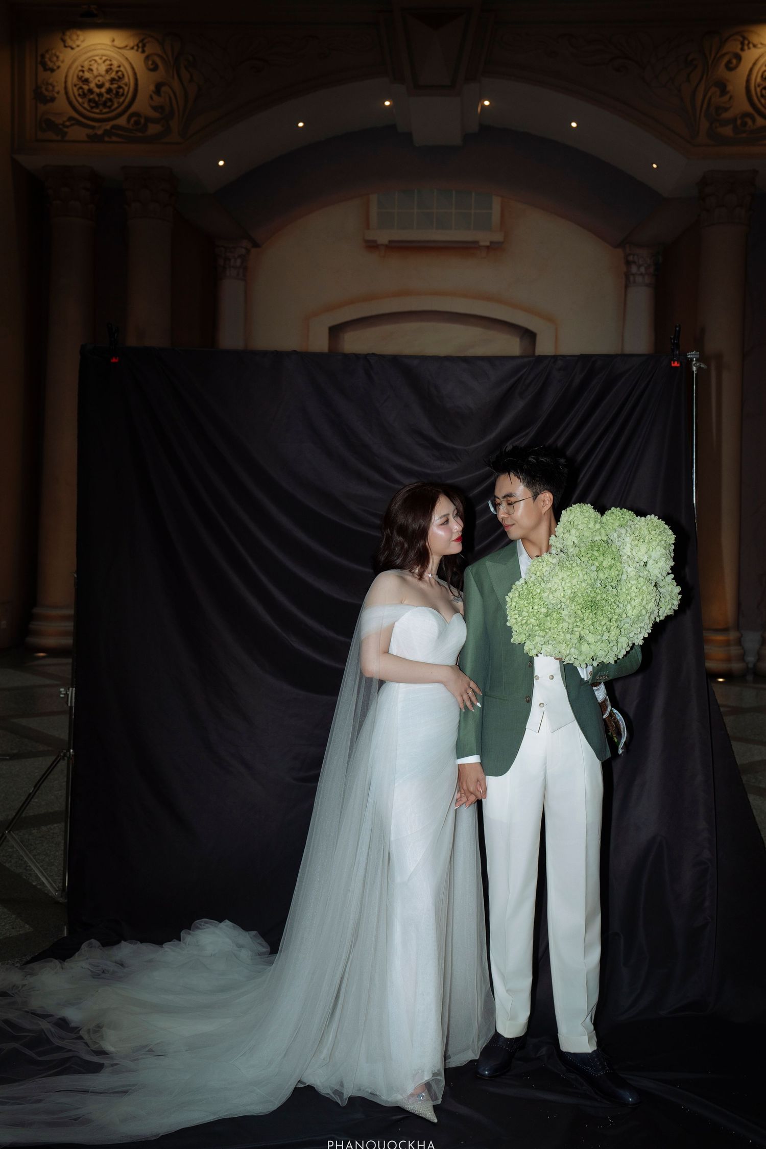 Bride and groom share intimate moment before black drape backdrop, groom holding large green bouquet.