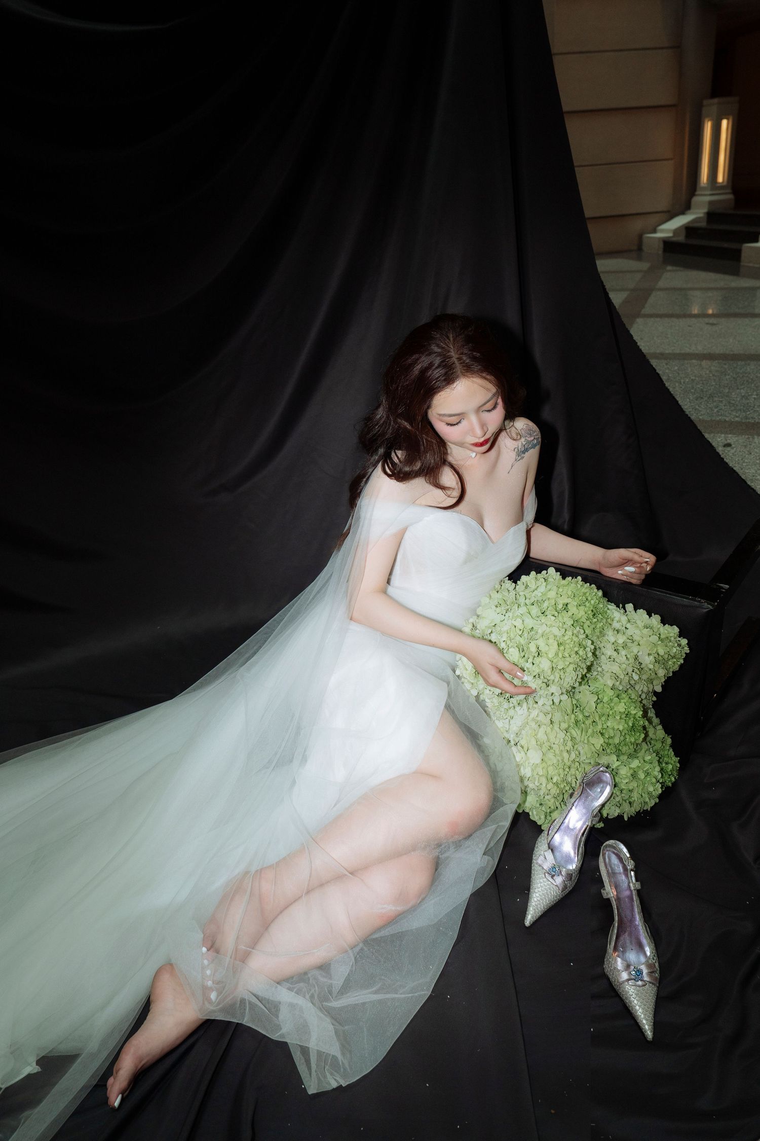 Bride in white tulle gown reclines on black fabric holding green hydrangea bouquet with glass heels.