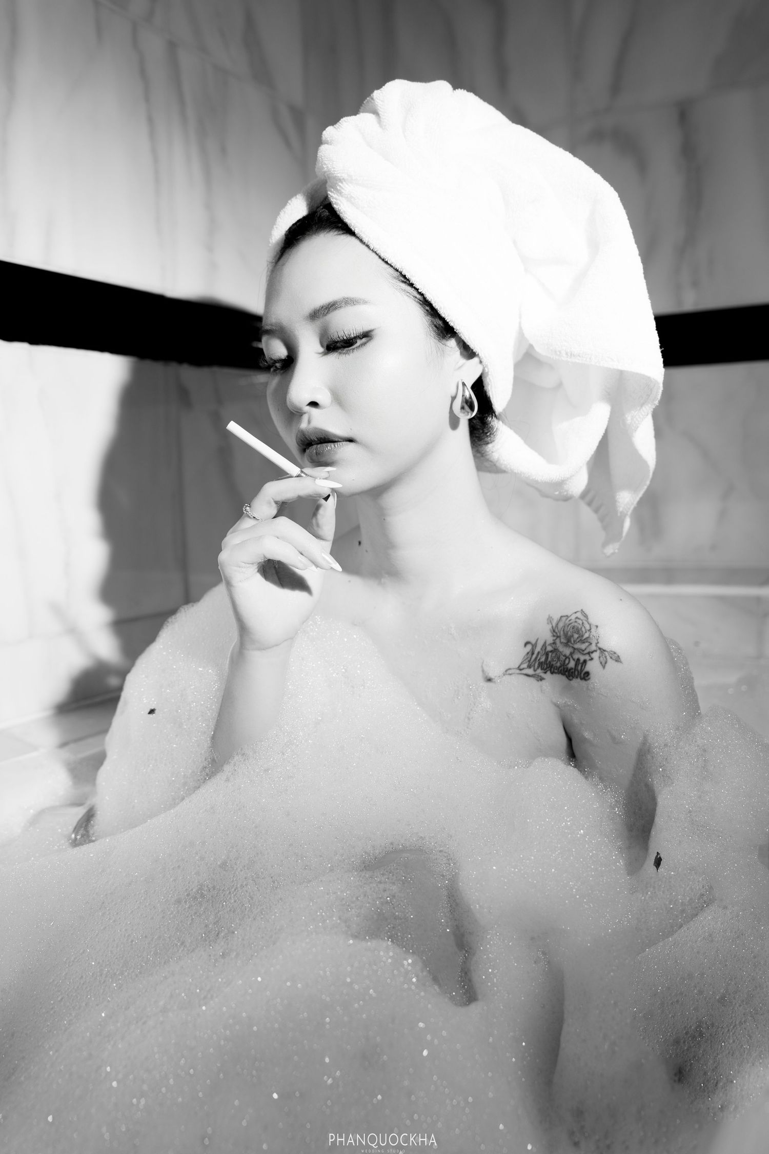 Black and white portrait of woman in bubble bath with towel turban holding a cigarette elegantly.