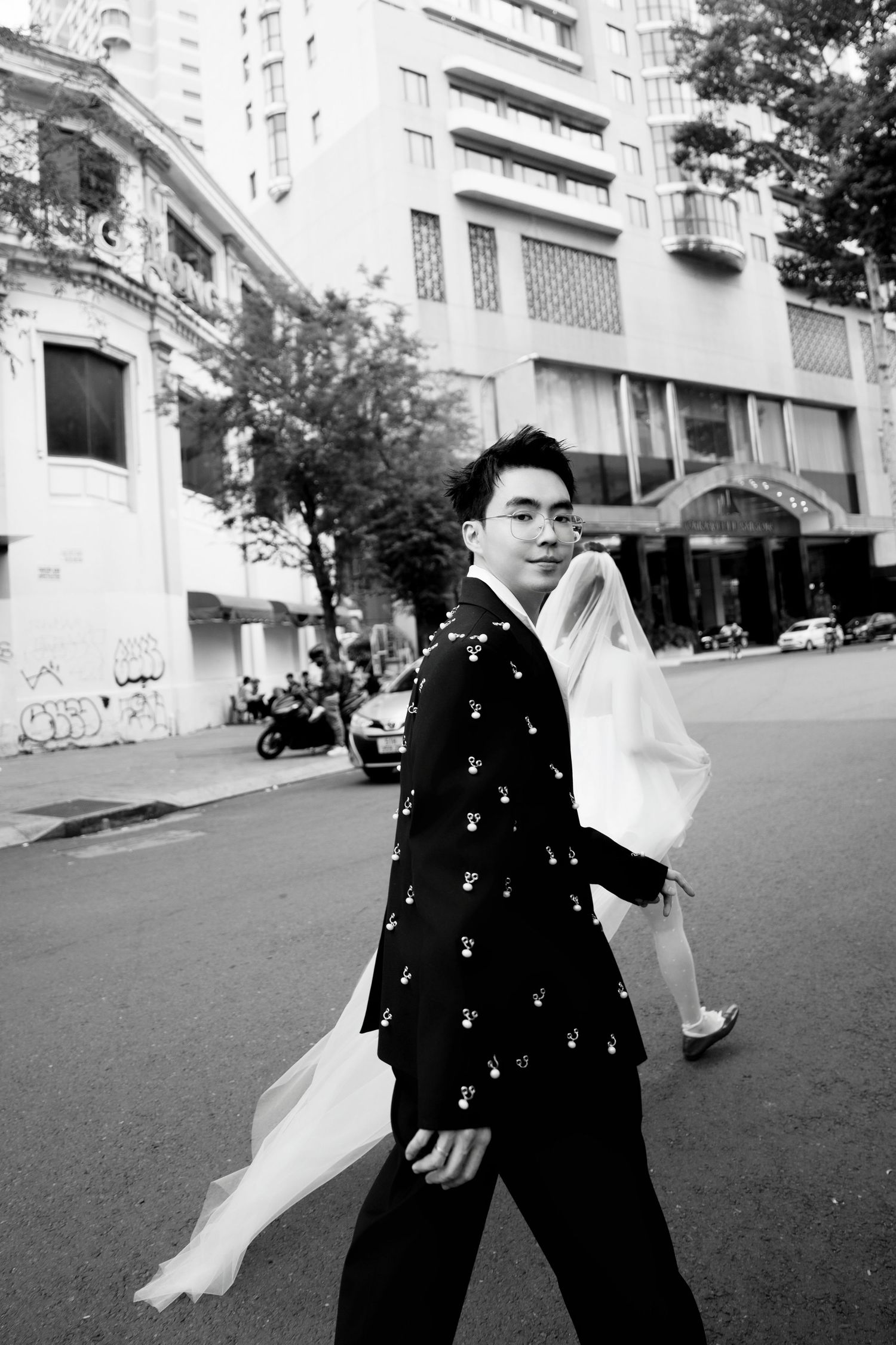Black and white photo of groom in embroidered suit leading bride across urban street with veil flowing.