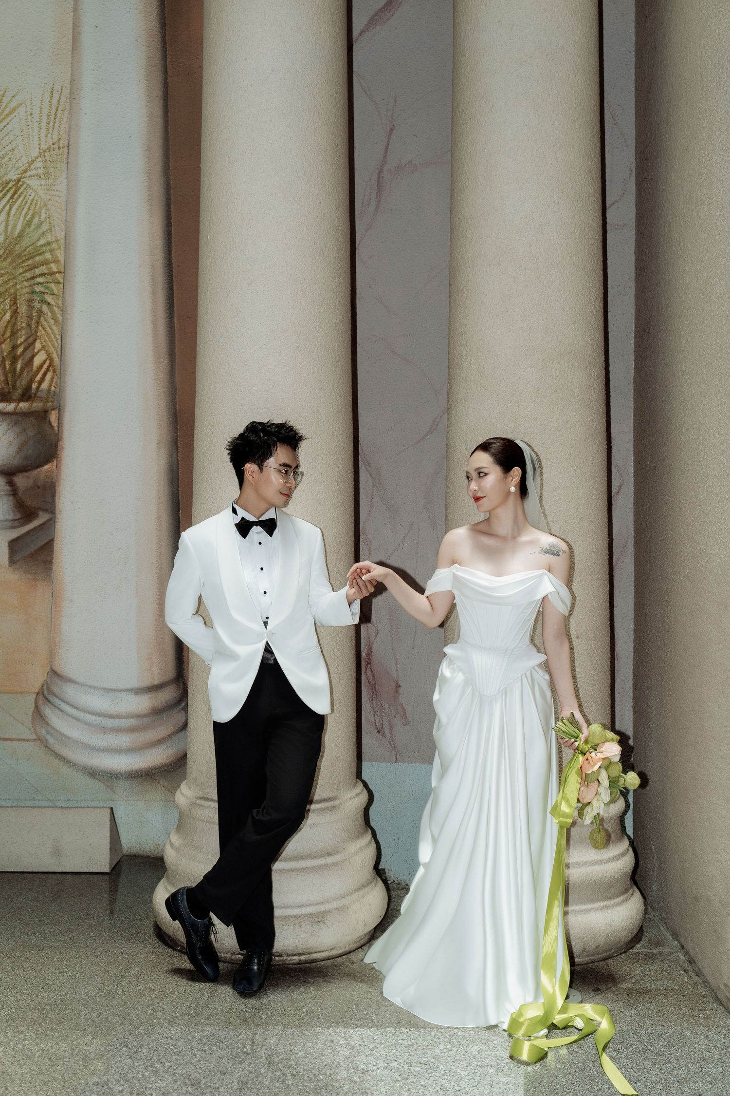 Elegant bride and groom posing by classical columns, groom in white tuxedo, bride in off-shoulder gown.