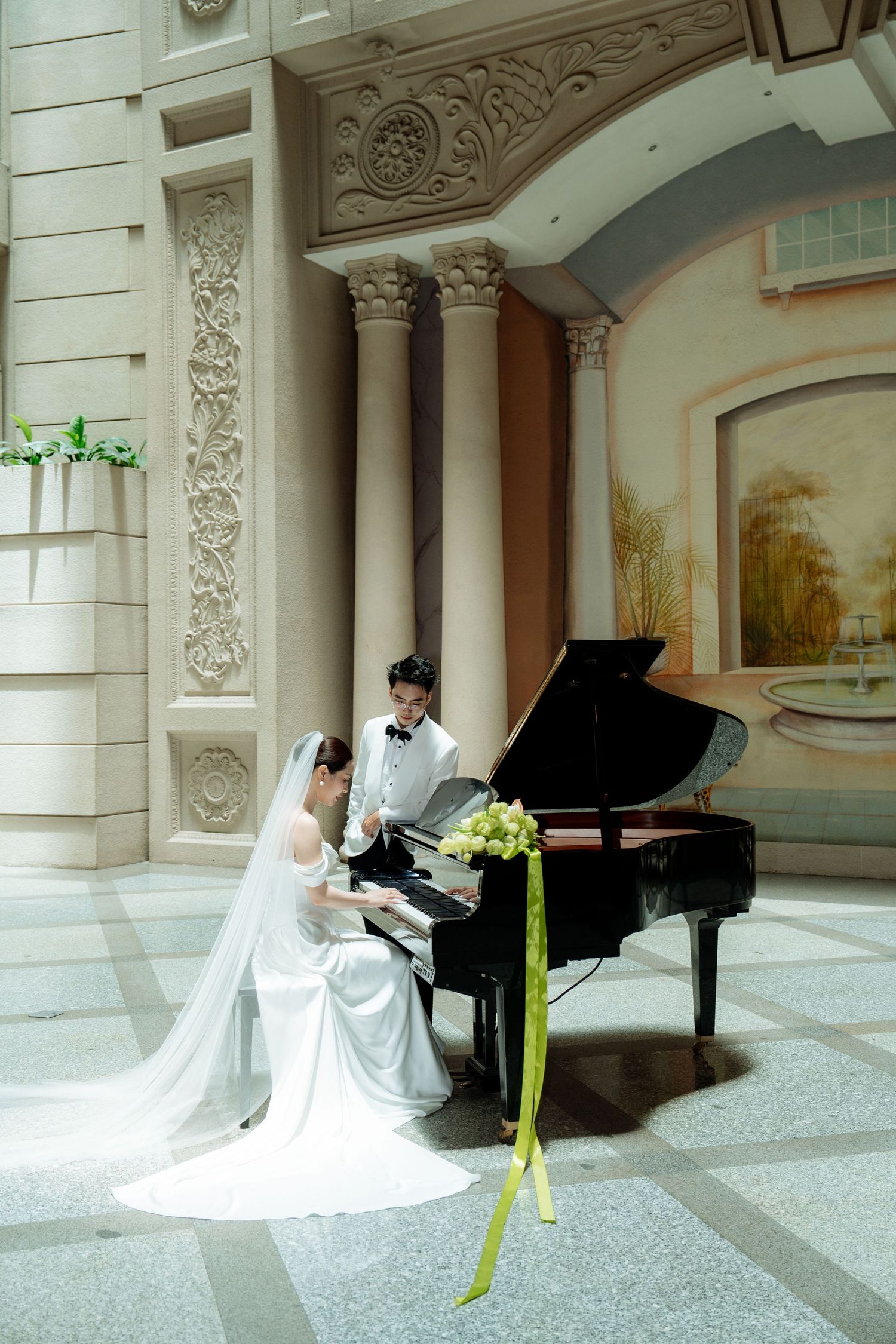 Bride in white gown playing grand piano with groom standing beside her in ornate classical interior.