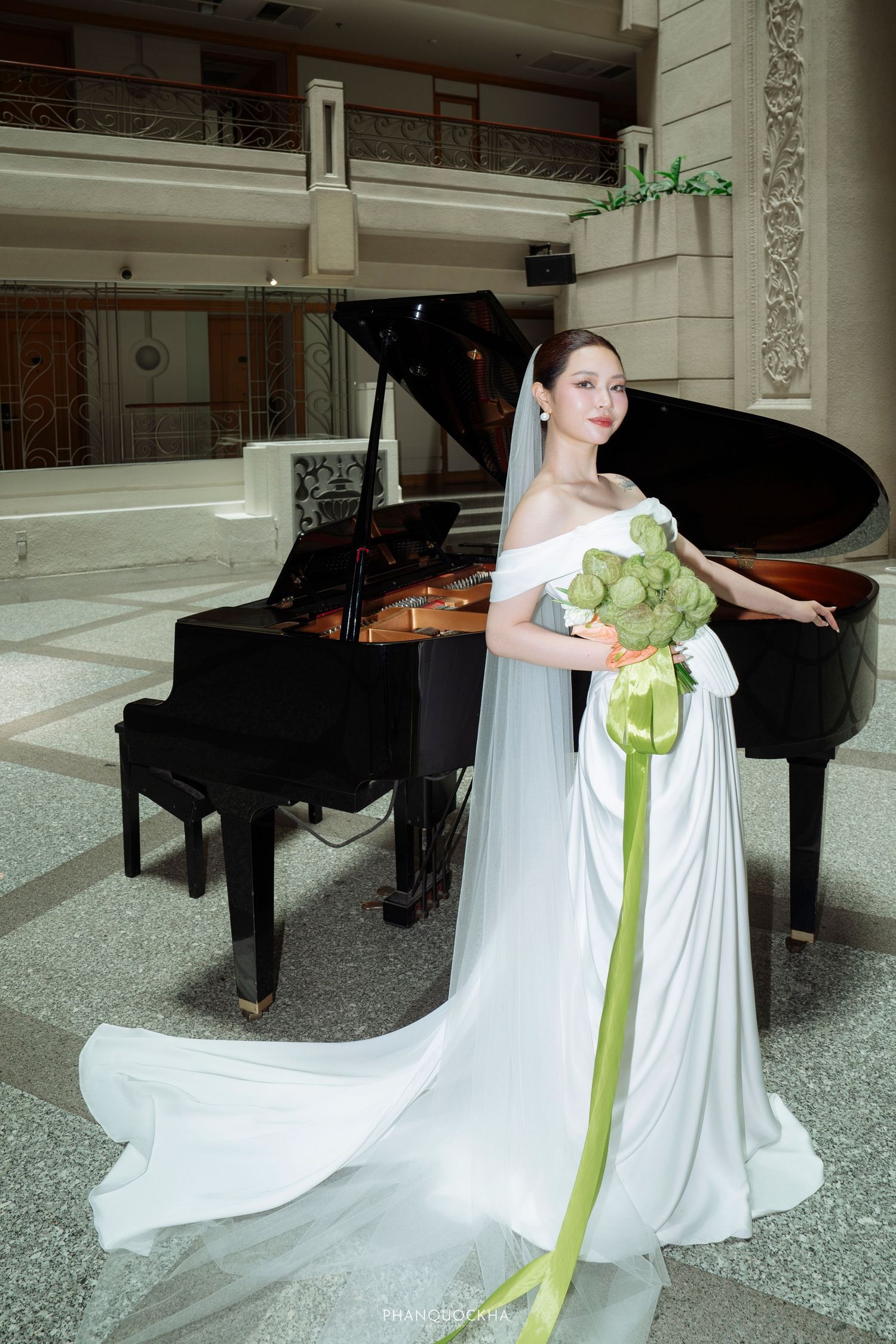Bride in elegant white gown with green floral bouquet poses gracefully beside a grand piano.