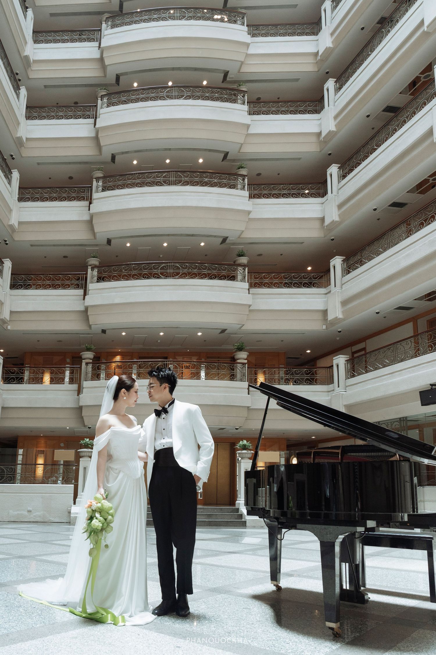 Bride and groom stand by a grand piano in an elegant multi-story hotel atrium with ornate balconies.