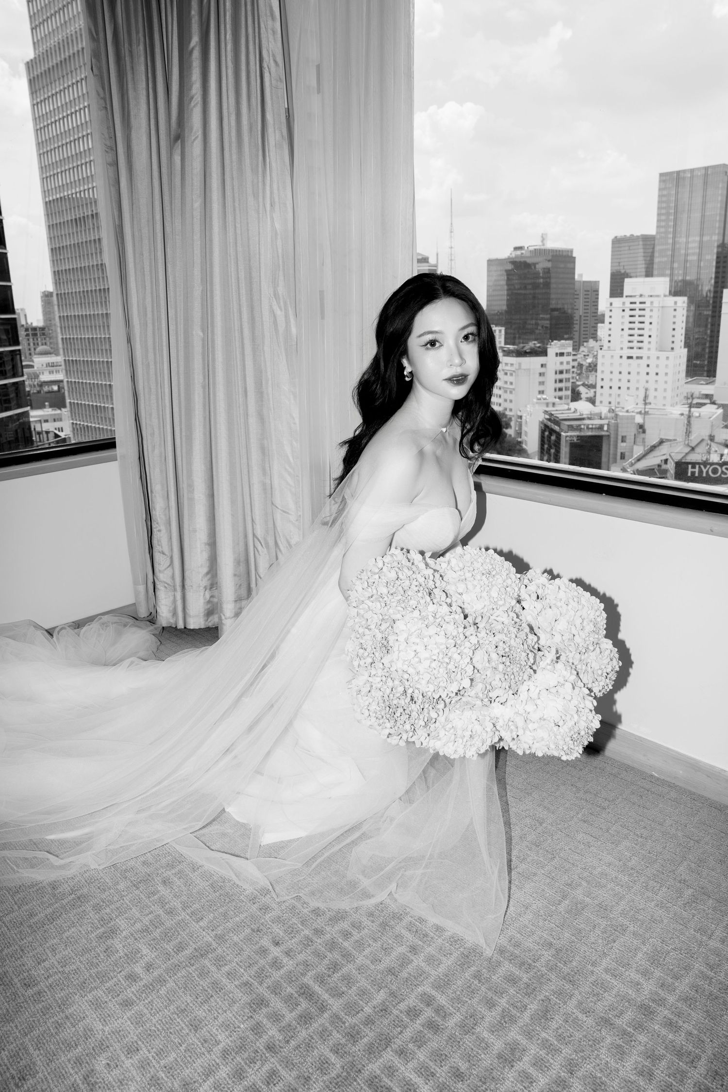 Black and white portrait of bride in gown holding large floral bouquet with city skyline background.