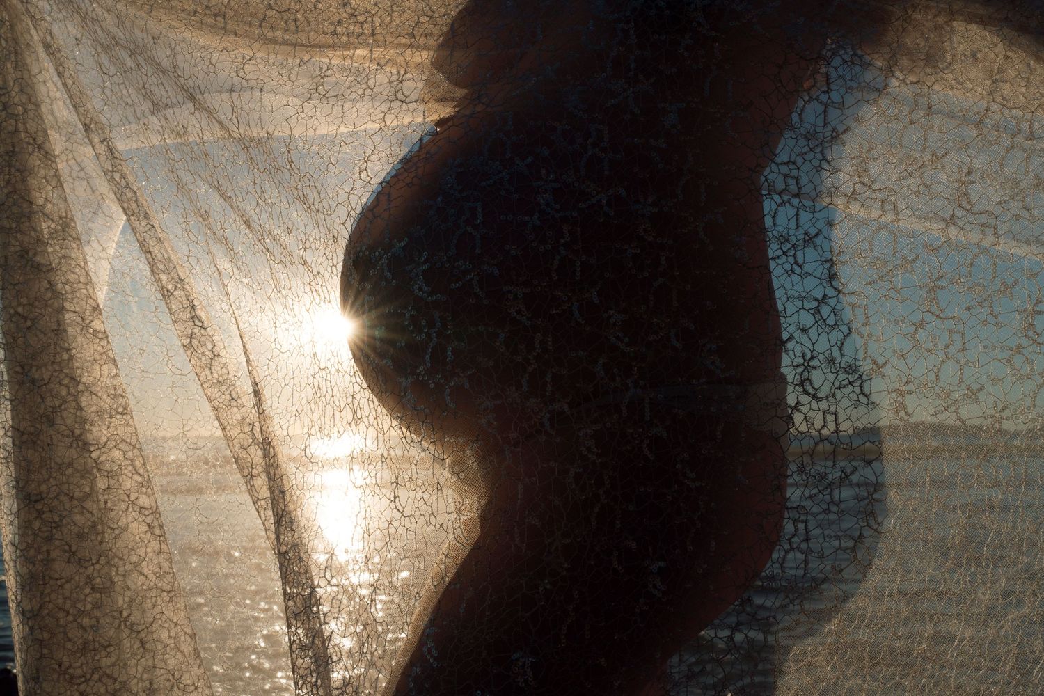 A silhouette of a pregnant woman's belly is illuminated through sheer fabric with sunlight creating a soft, ethereal glow.