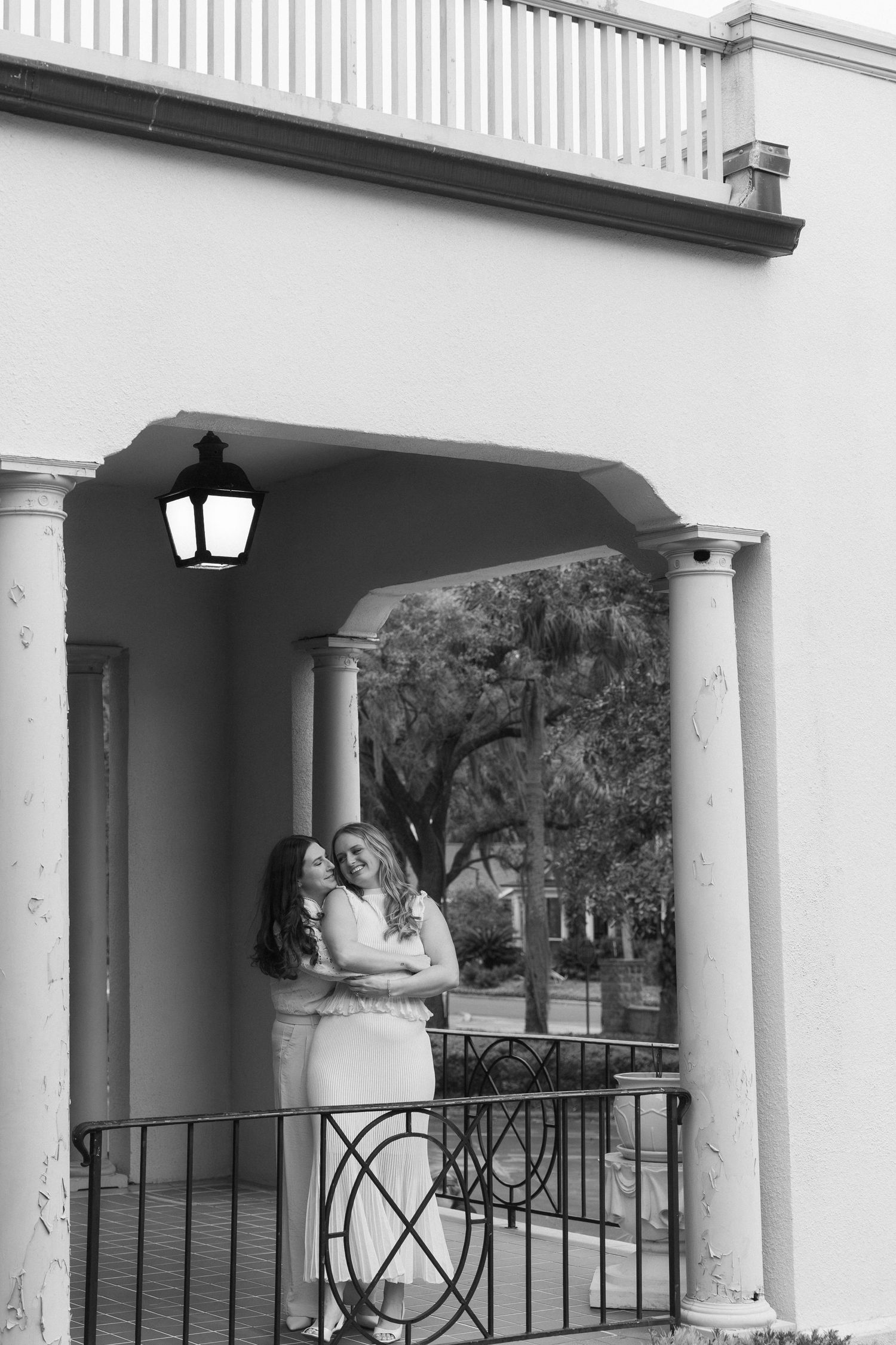 Two women in white dresses embrace on a classic columned porch in this elegant black and white portrait.