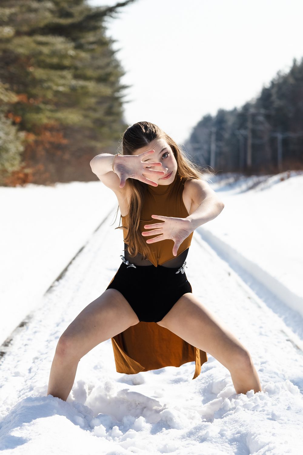 A person in winter attire poses playfully in deep snow on a tree-lined path during a bright winter day.