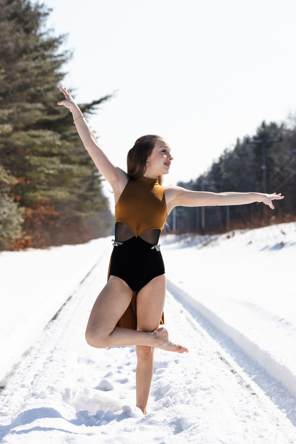 A dancer performs a graceful leap sequence in the snow wearing a black leotard against an evergreen forest backdrop.