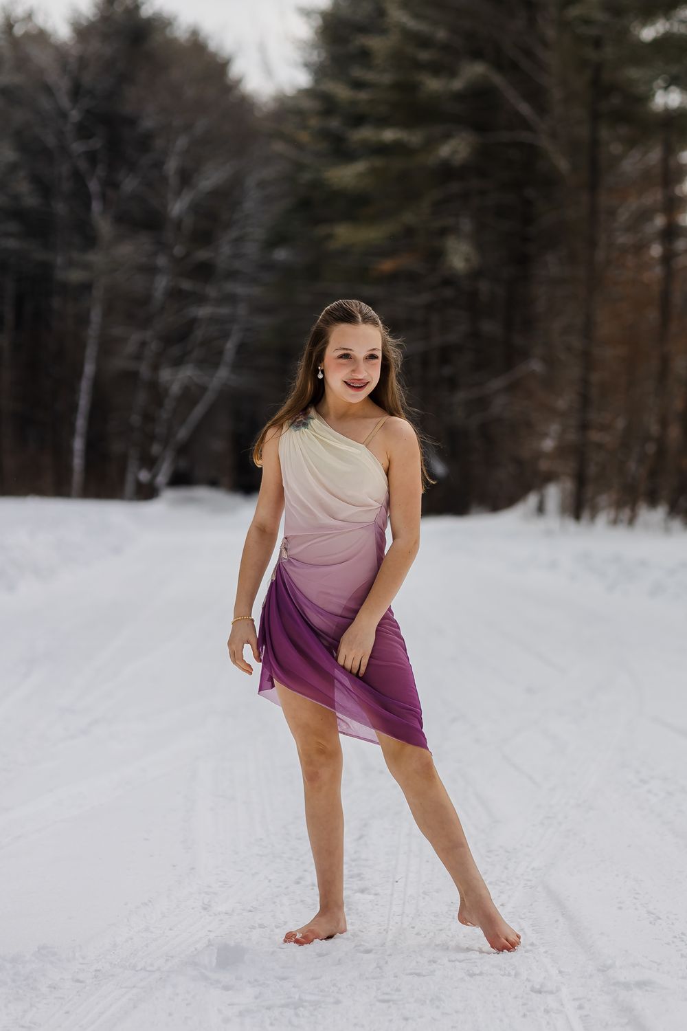 A person in an ombre dress performs a graceful dance pose on a snowy winter landscape with evergreen trees.