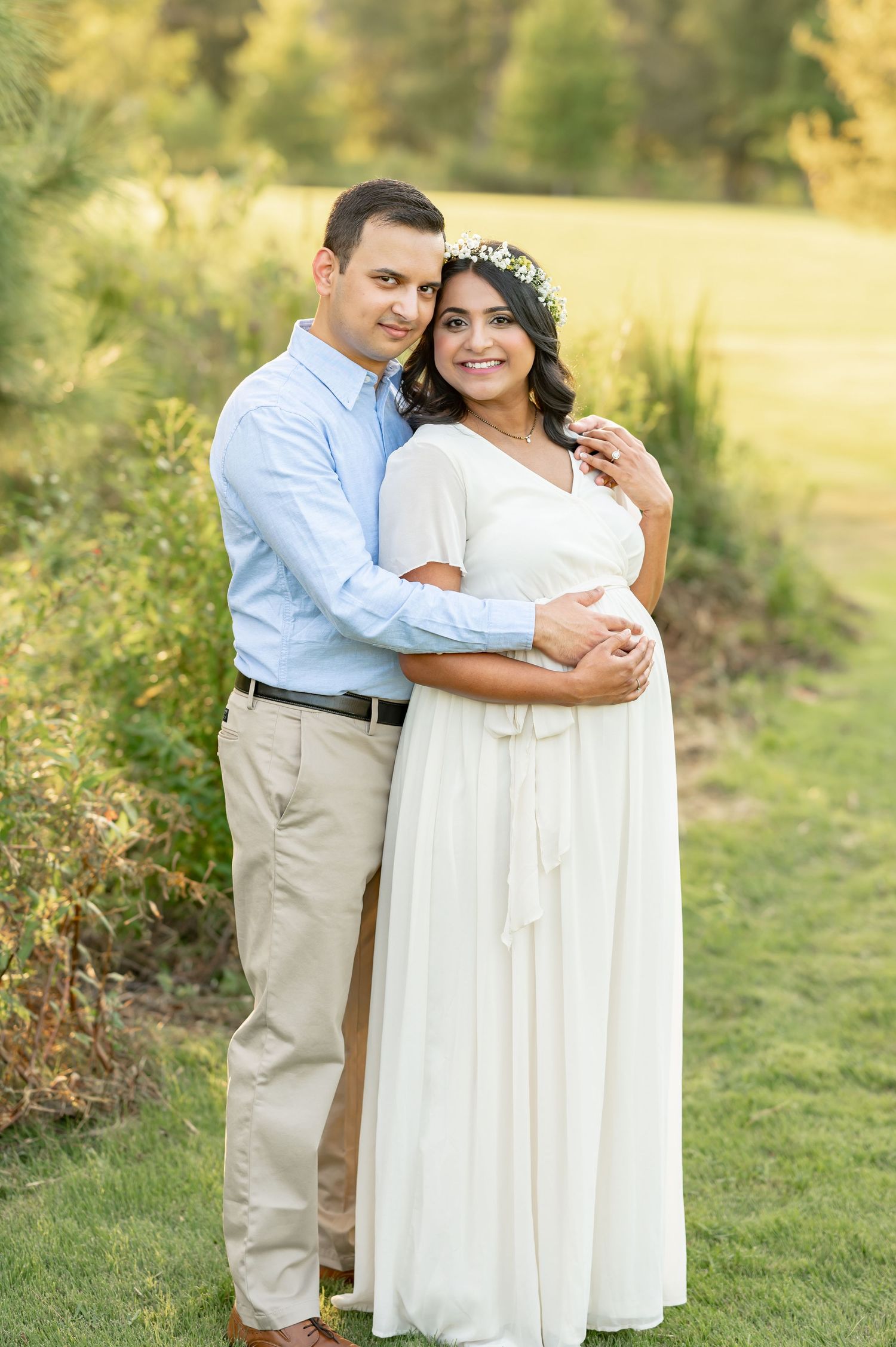A couple embraces outdoors in a natural setting, wearing a white flowing dress and light blue shirt against a sunny backdrop maternity