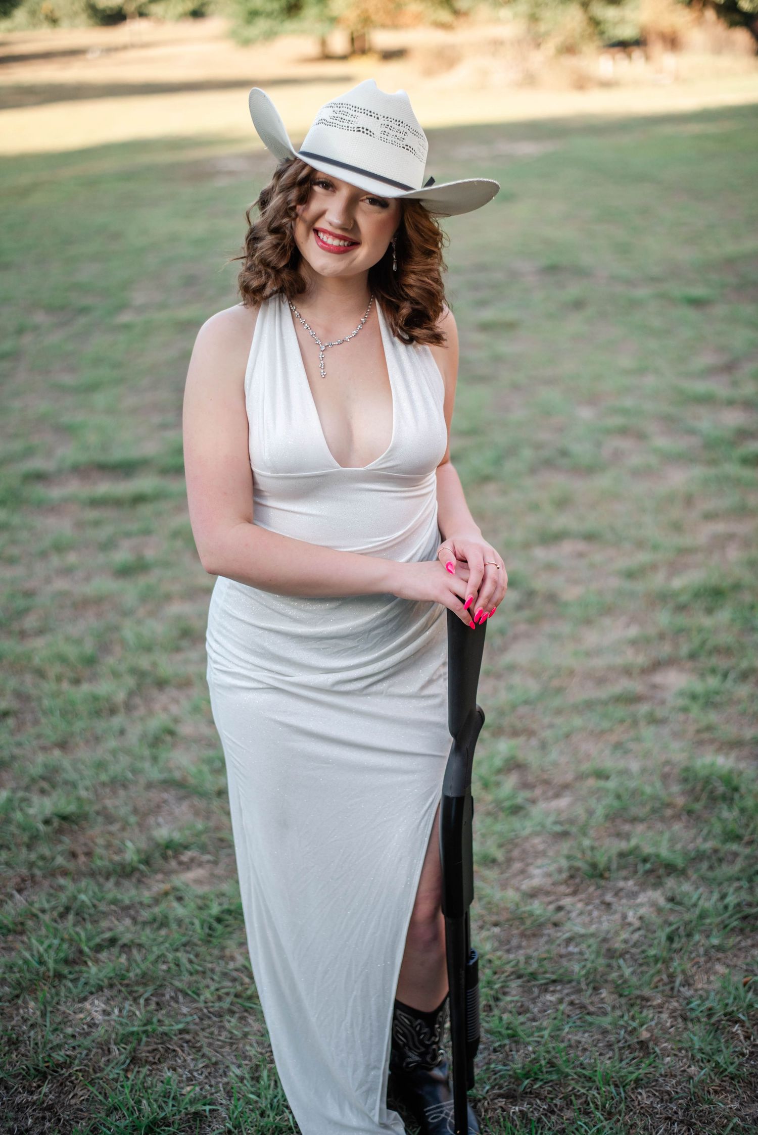Woman in white halter dress and cowboy hat poses outdoors holding a rifle, wearing black cowboy boots.