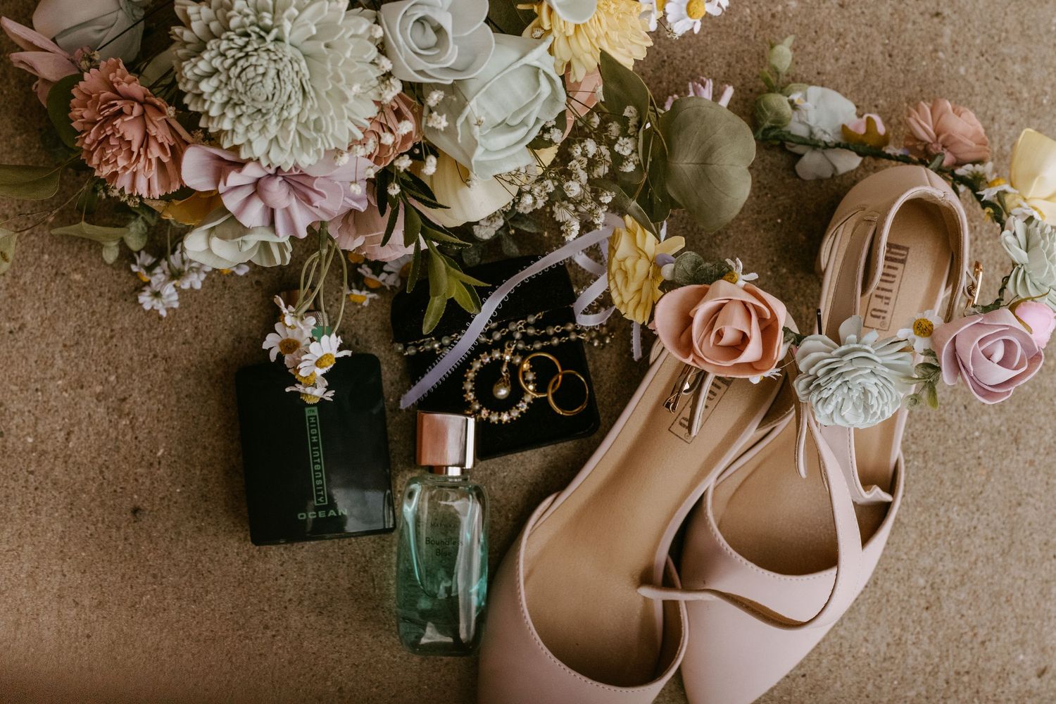 Bridal flat shoes with floral detail beside perfume bottles, gold jewelry, and a pastel flower bouquet on a rustic surface.