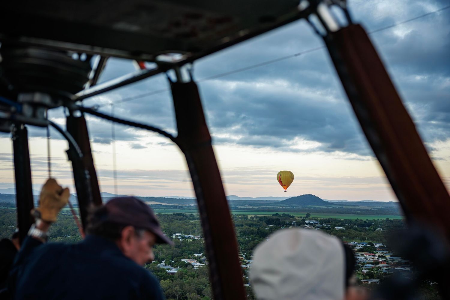 View from hot air balloon basket showing another yellow balloon floating over green landscape under cloudy sky. Cairns, Atherton