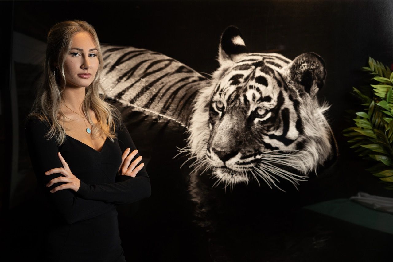 Woman in black dress poses confidently beside a large black and white tiger painting in a dark studio setting.
