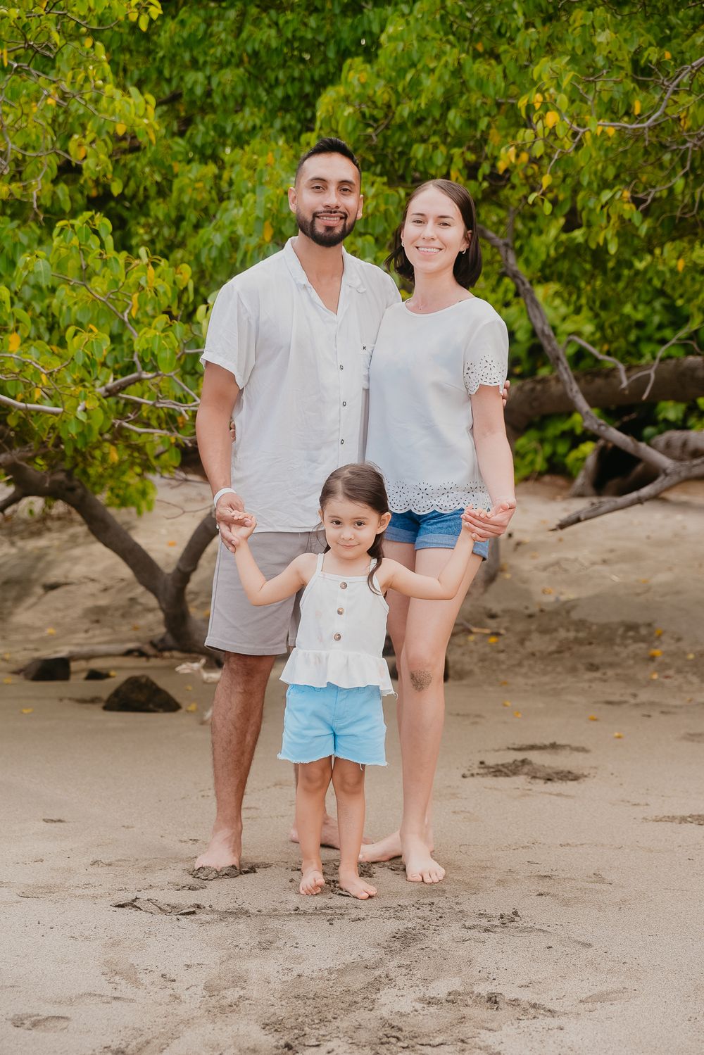 Family portrait session at a scenic beach with lush green trees and sand, capturing casual moments together.