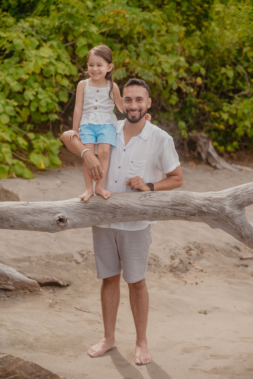 Two people share a tender moment while sitting on driftwood at a sandy beach with lush greenery in the background.