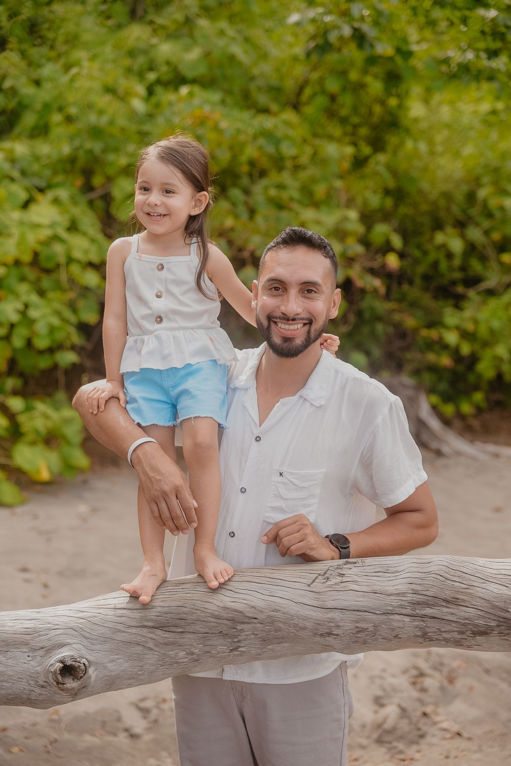 Family enjoying a playful moment together on a fallen tree trunk at the beach with lush greenery in background.
