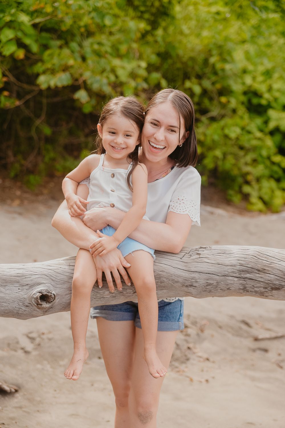A tender moment shared between two people sitting on a fallen log at the beach surrounded by green foliage.