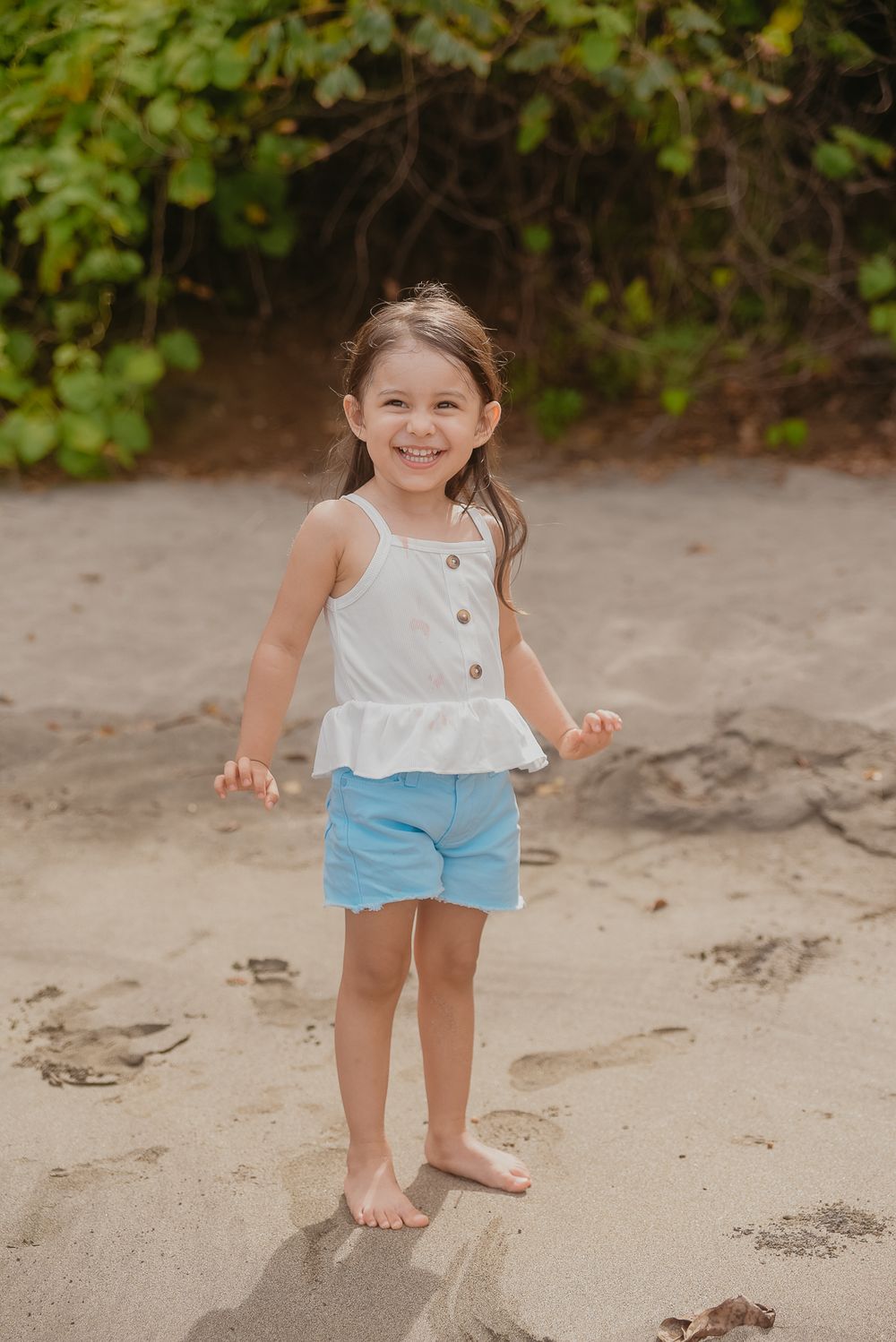 A child in a white top and blue shorts plays on a sandy beach with green foliage in the background.