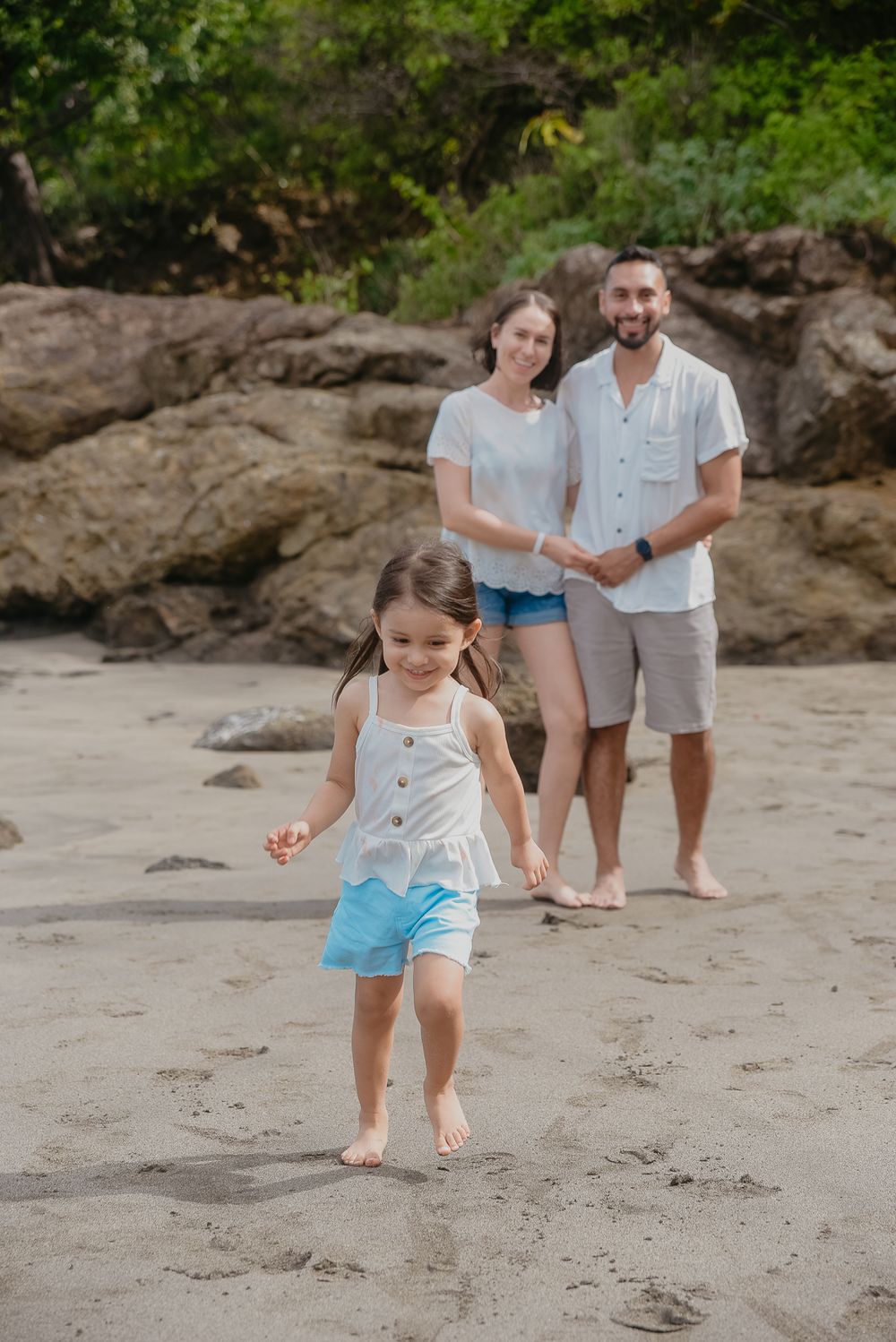 Family plays and jumps together on a sandy tropical beach with rocky outcrops and lush greenery in background.