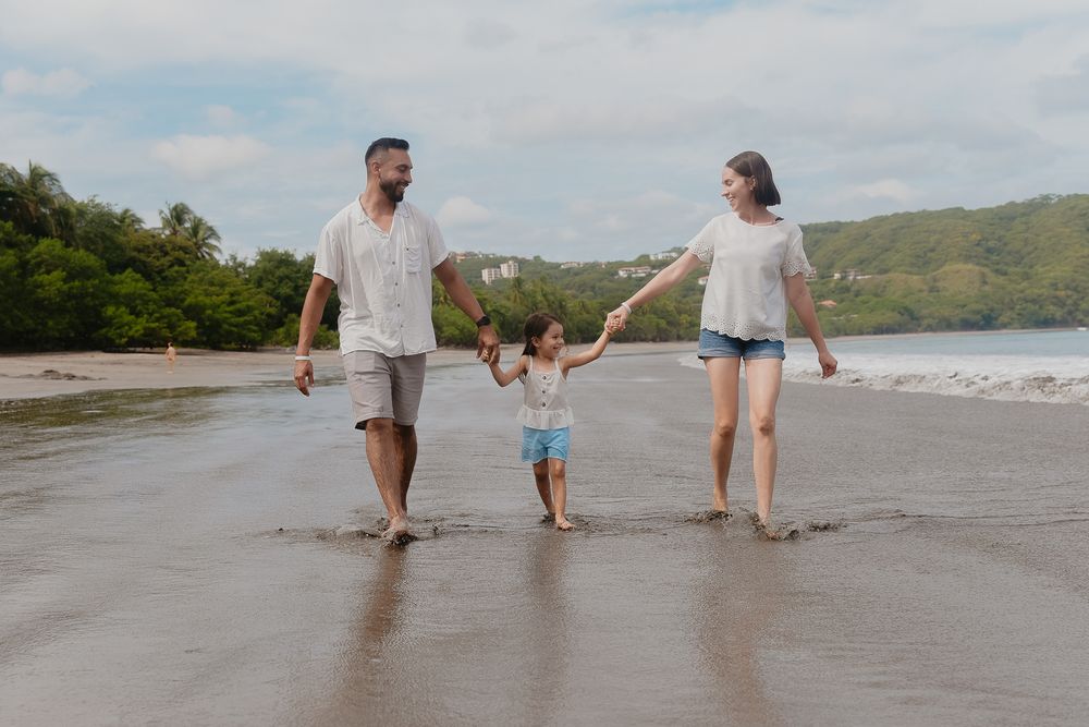 Family members walk along the shoreline of a beach with mountains visible in the distance.