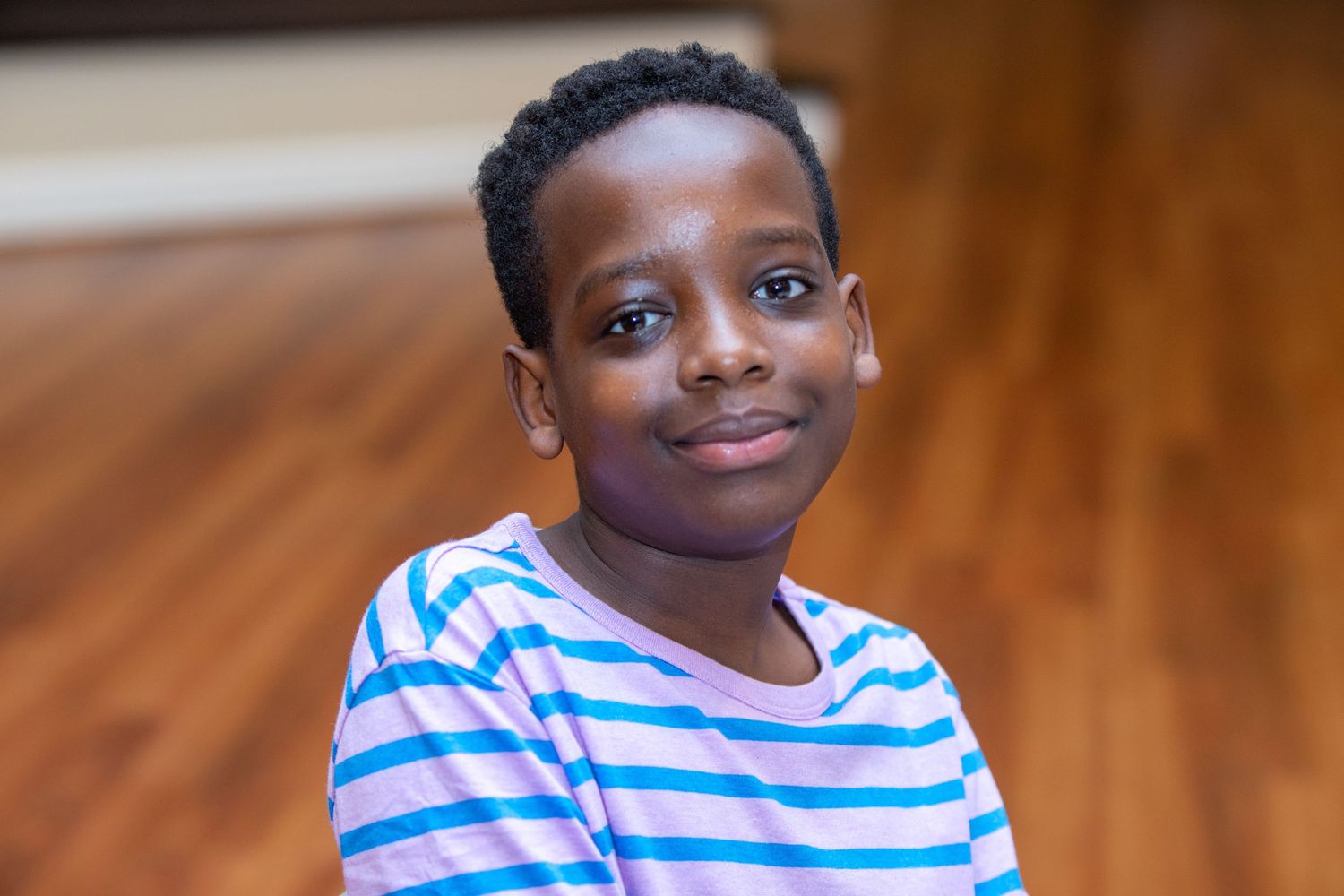 A smiling youth in a striped shirt poses for a portrait on hardwood floors.