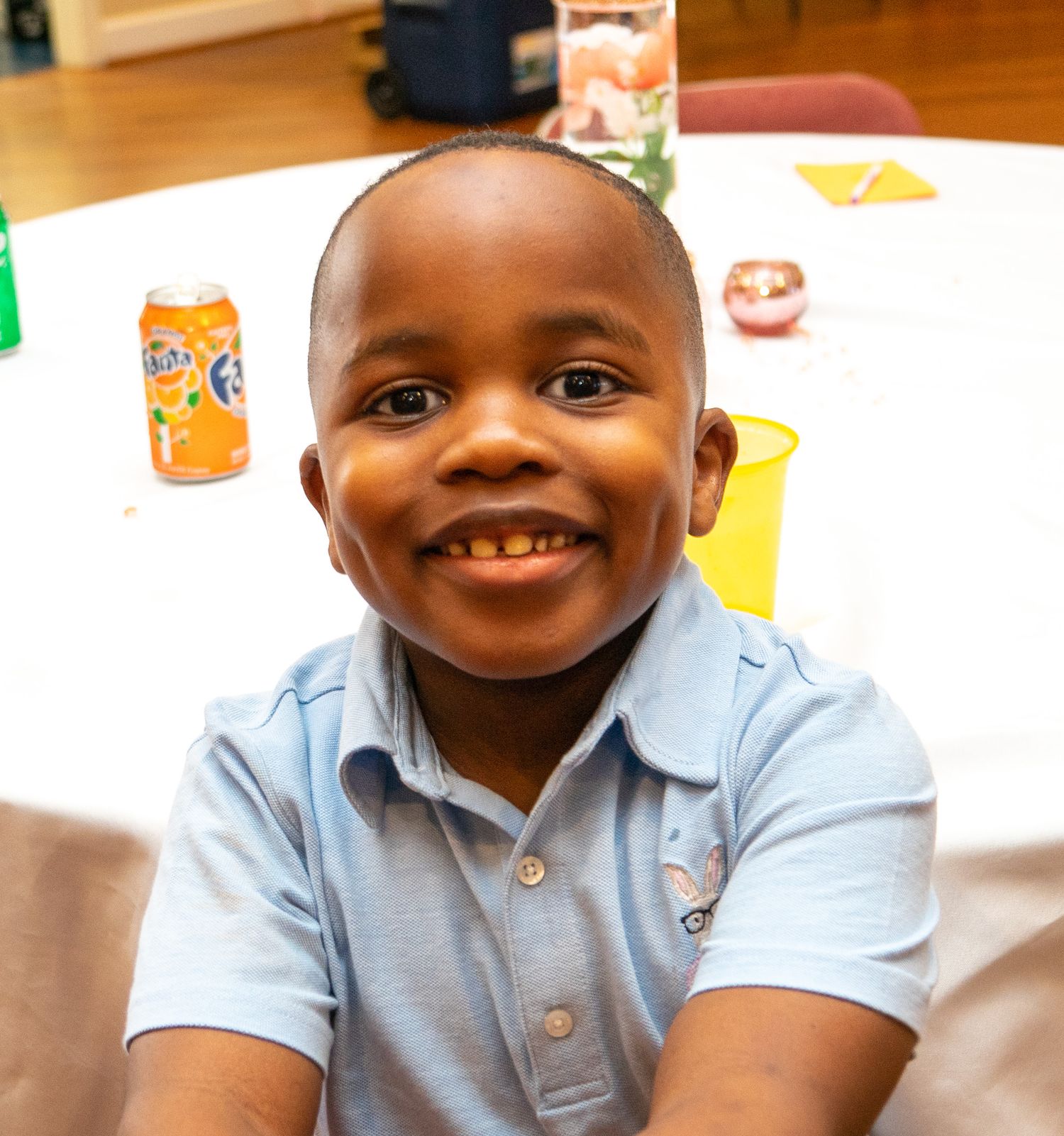 A child in a light blue polo shirt smiles brightly at a table with drinks in the background.