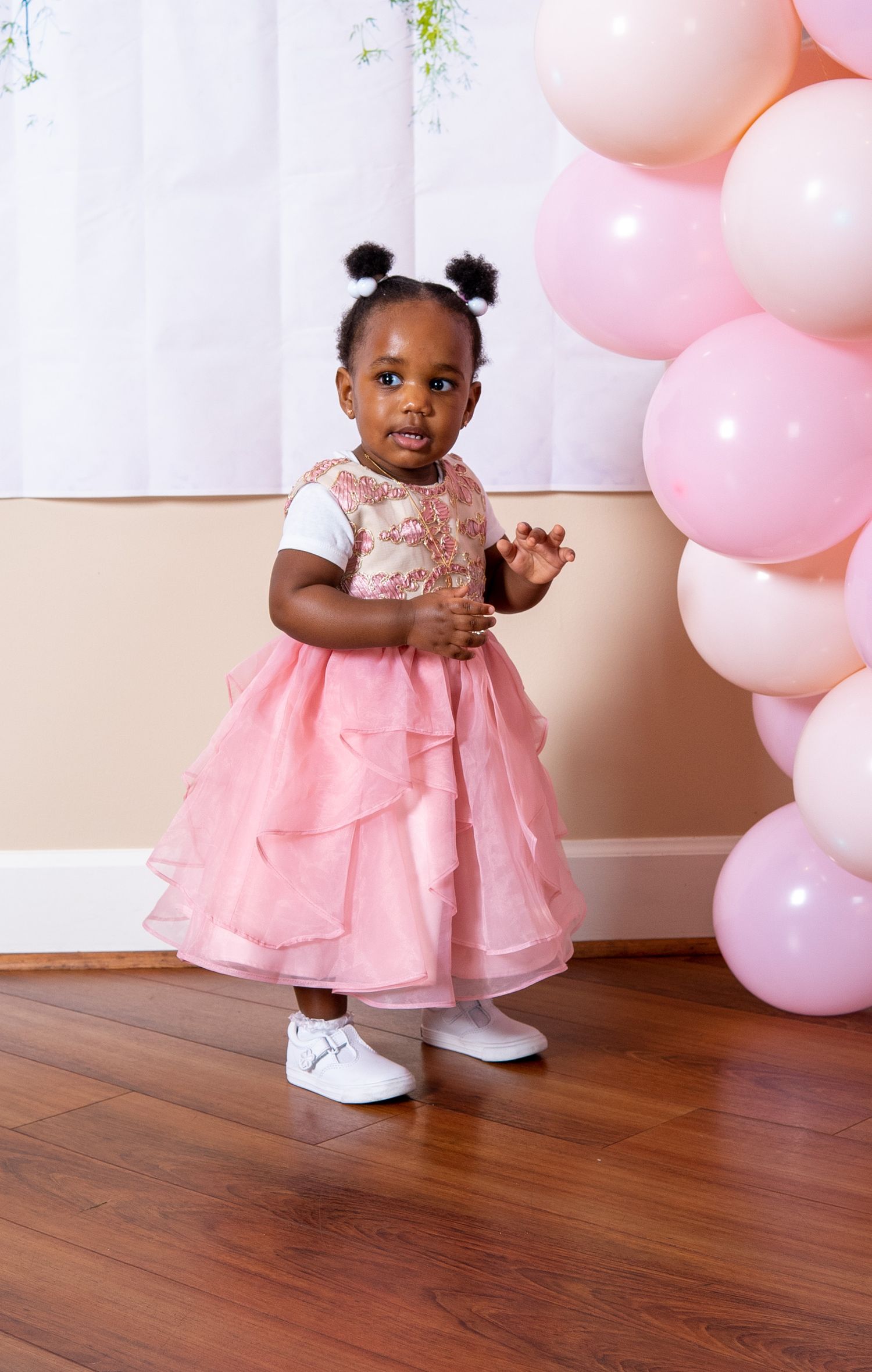 A toddler in a pink tulle dress poses in front of pink balloons at a party.
