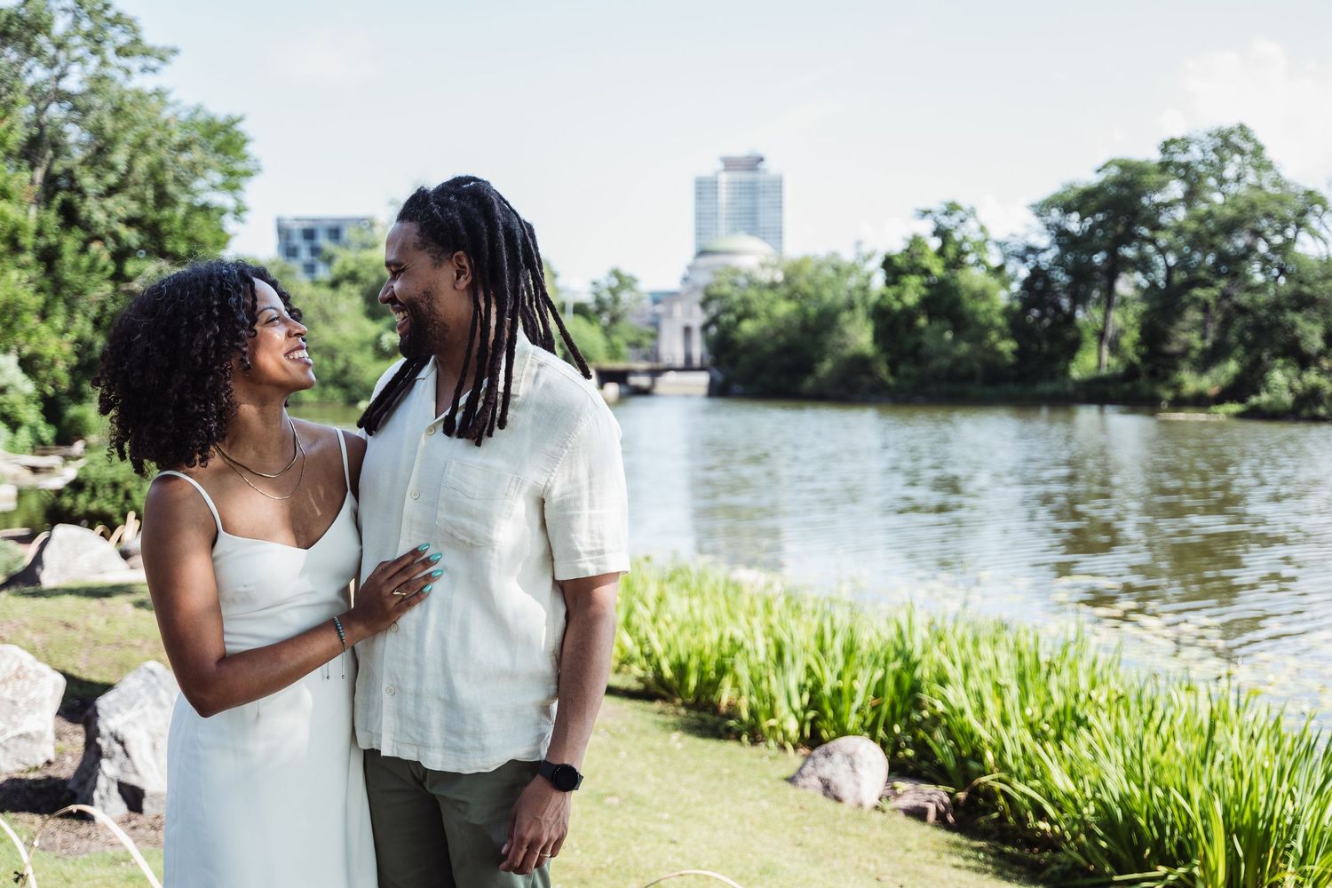 A romantic couple stands by a serene lake with lush greenery and city buildings in the background.