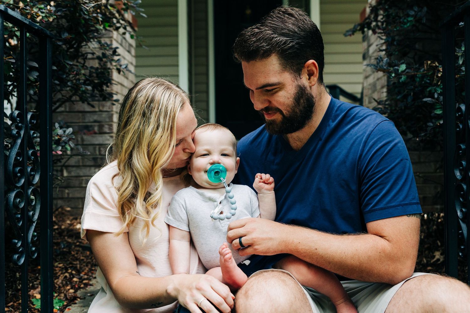 A family sits together on their front porch steps sharing a tender moment with their baby wearing a teal pacifier.
