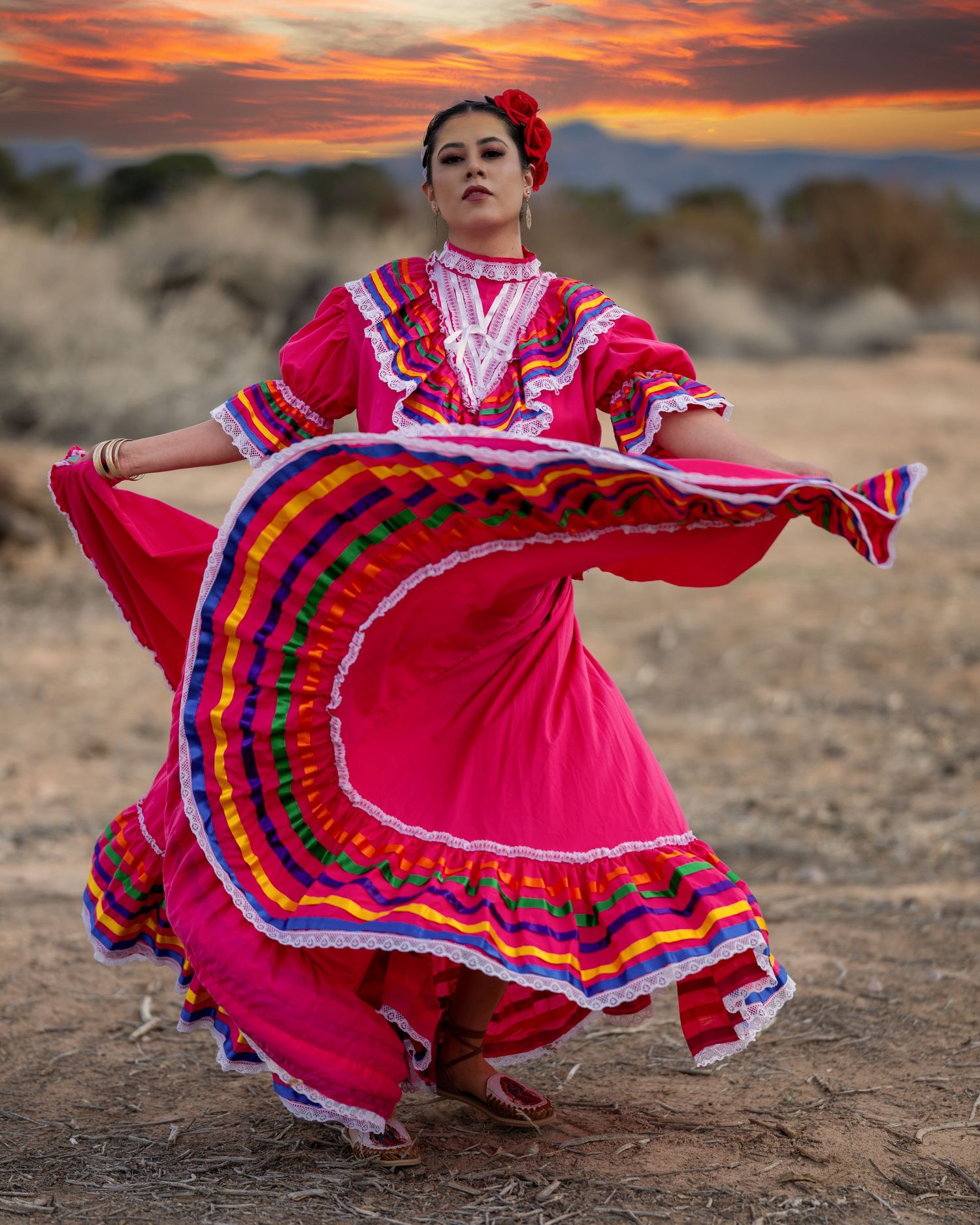 Woman in a vibrant pink traditional folklórico dress with multicolored ribbon trim twirls gracefully in a desert landscape at sunset.