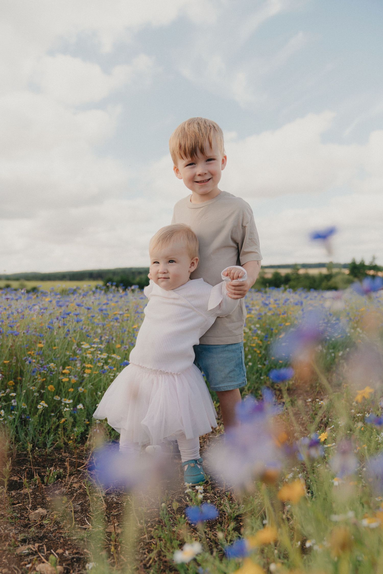 Two young children smiling in a colorful wildflower field on a cloudy day.