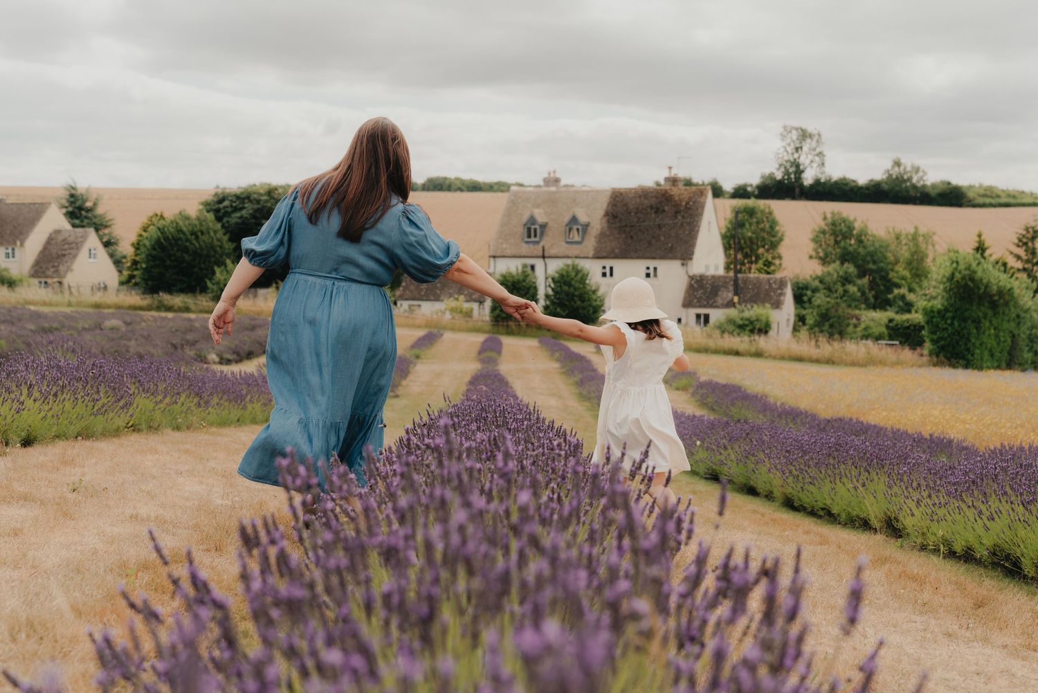 A mother and child walk hand-in-hand through a blooming lavender field near a countryside farmhouse.