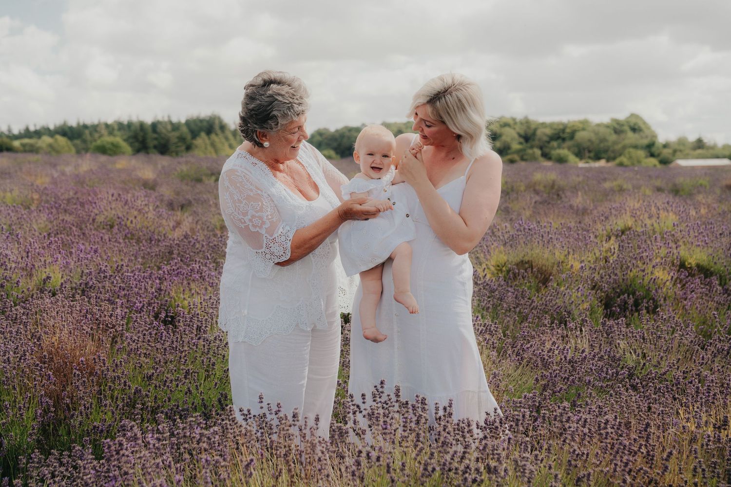 Three generations of women in white dresses stand in a blooming lavender field, lovingly admiring a baby.