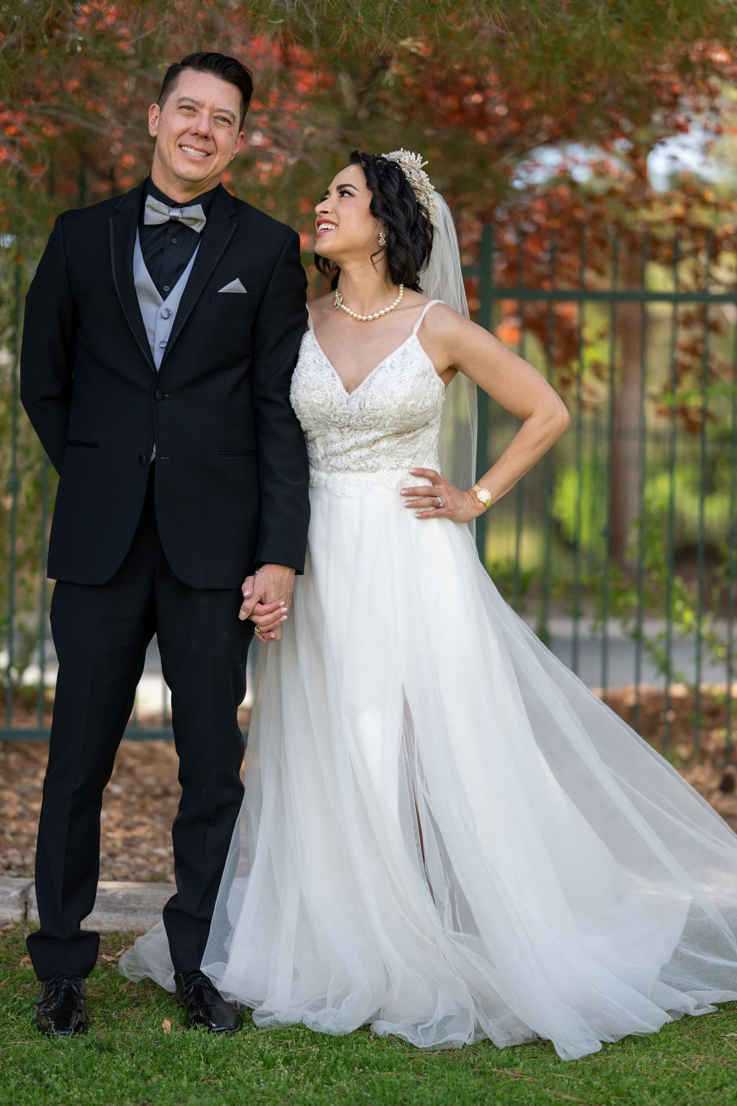 Elegant outdoor wedding portrait of a bride and groom holding hands, smiling together beneath autumn-colored trees; the groom wears a black