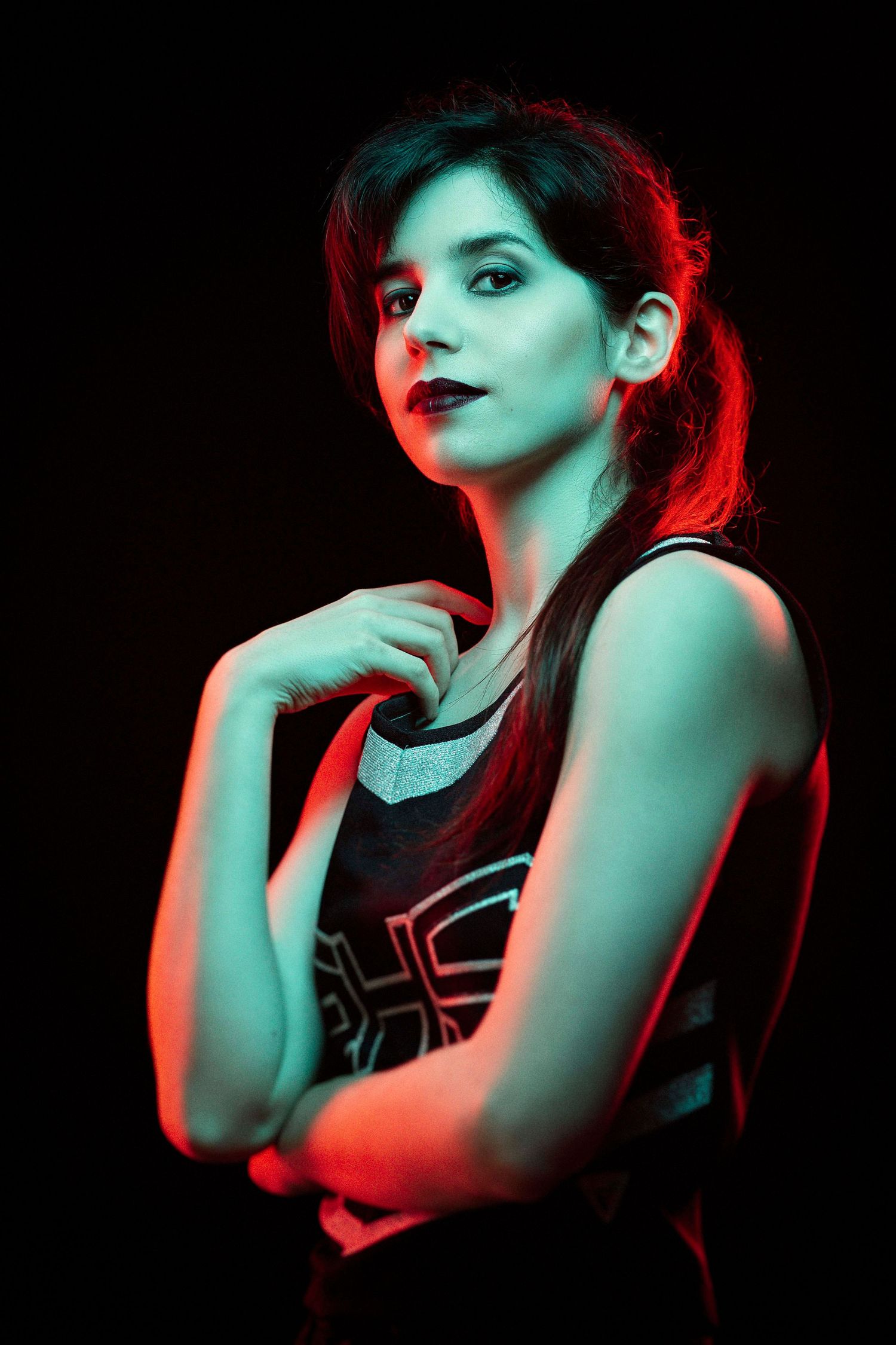 Young woman in sleeveless top poses confidently under dramatic red and teal studio lighting against black background.