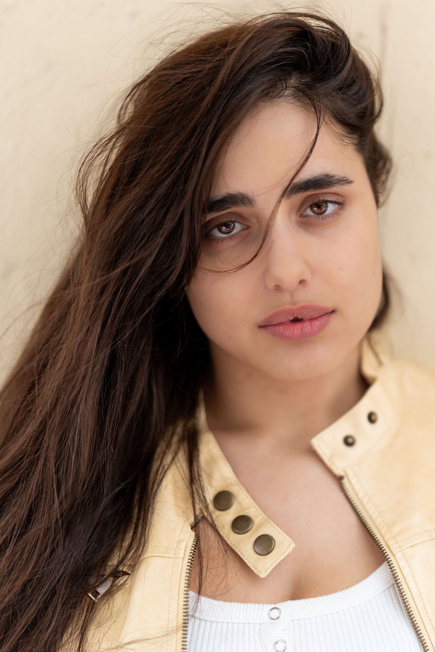 Young woman with long dark hair wearing a yellow leather jacket, posing against a beige wall.