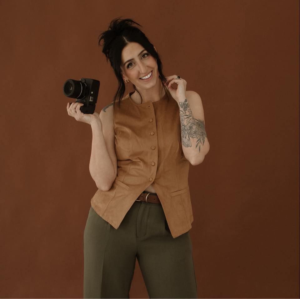 Woman in brown vest holding camera, smiling, with floral arm tattoo, against warm brown background.