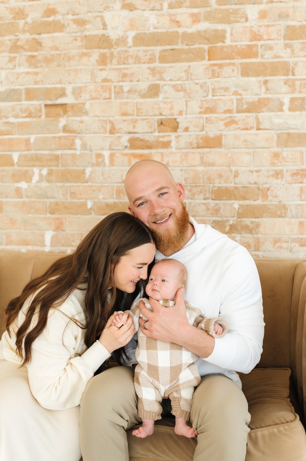 A family shares tender moments on a beige couch against a rustic brick wall in a series of lifestyle portraits.