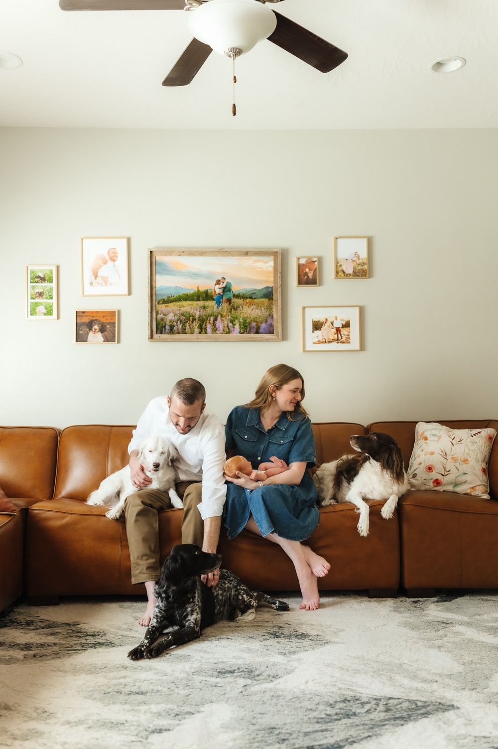 A couple sits on a brown leather couch with their dog in a cozy living room with framed artwork on the walls.