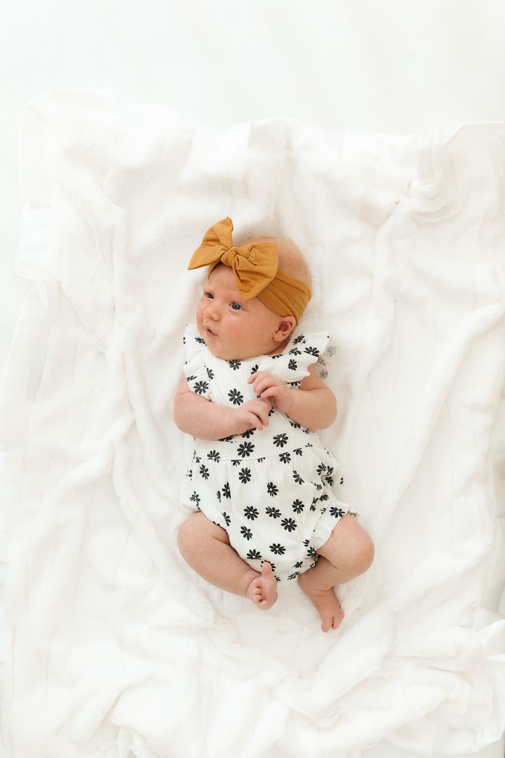 A baby wearing a floral onesie and mustard yellow bow headband lies peacefully on white bedding.