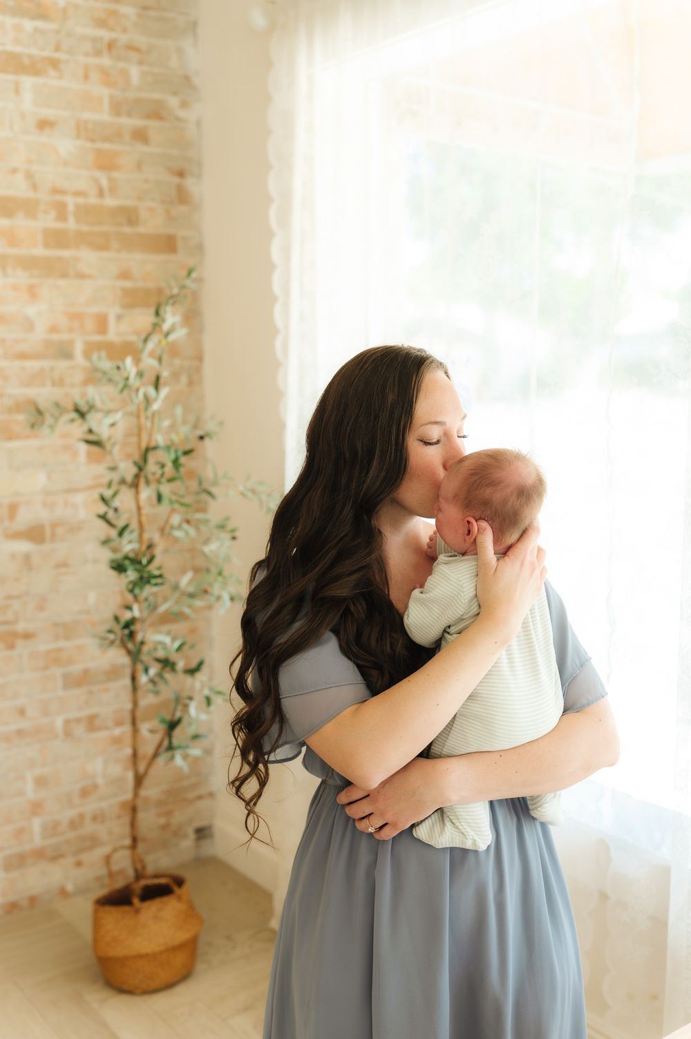 A tender moment between parent and baby in a sunlit room with exposed brick walls and a potted plant.
