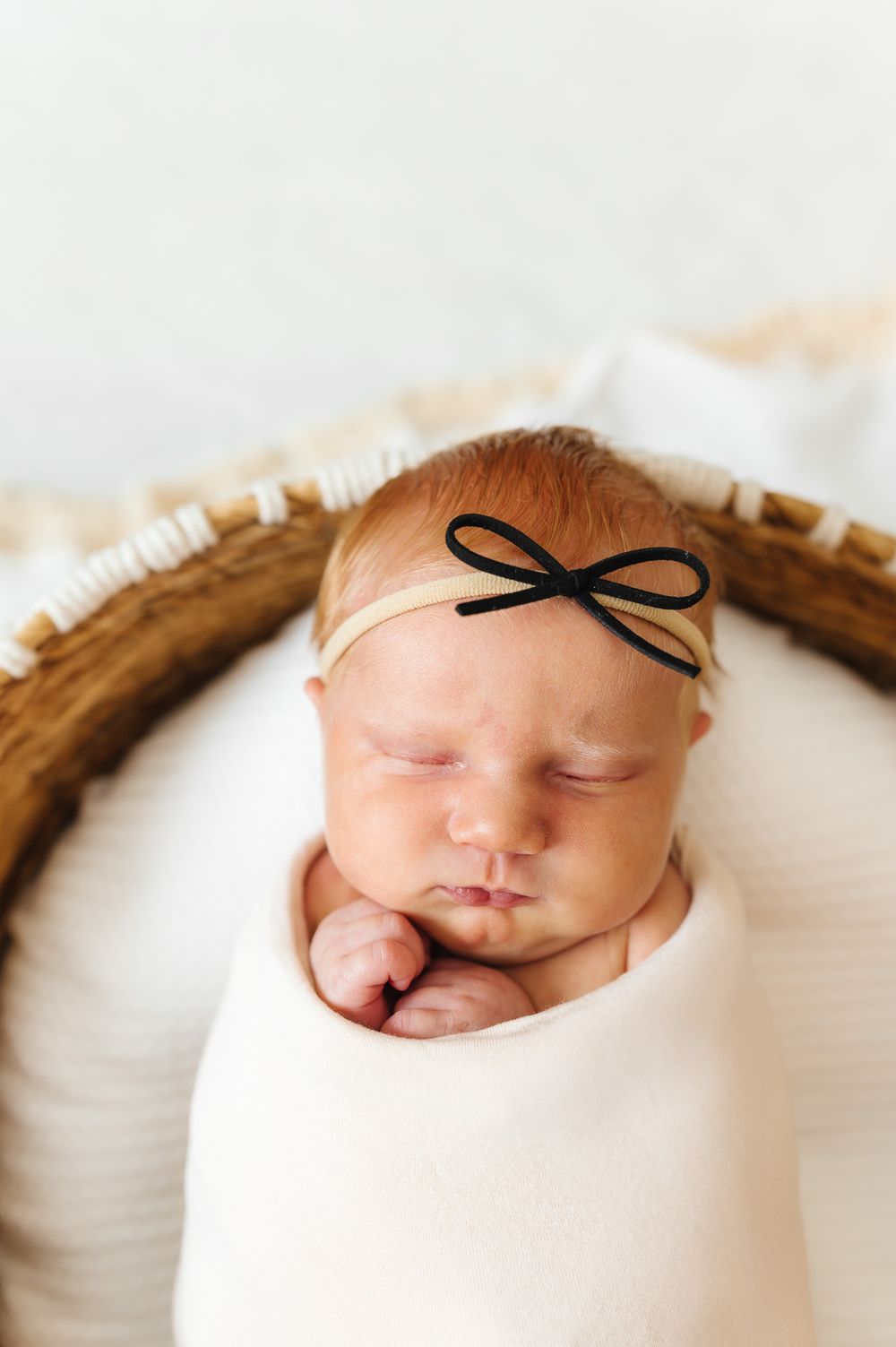 A sleeping newborn baby swaddled in white fabric wearing a delicate black bow headband.
