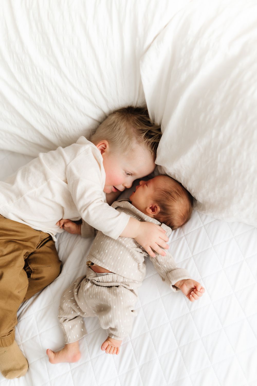 Two siblings share a tender moment lying together on white bedding, creating a heartwarming scene.