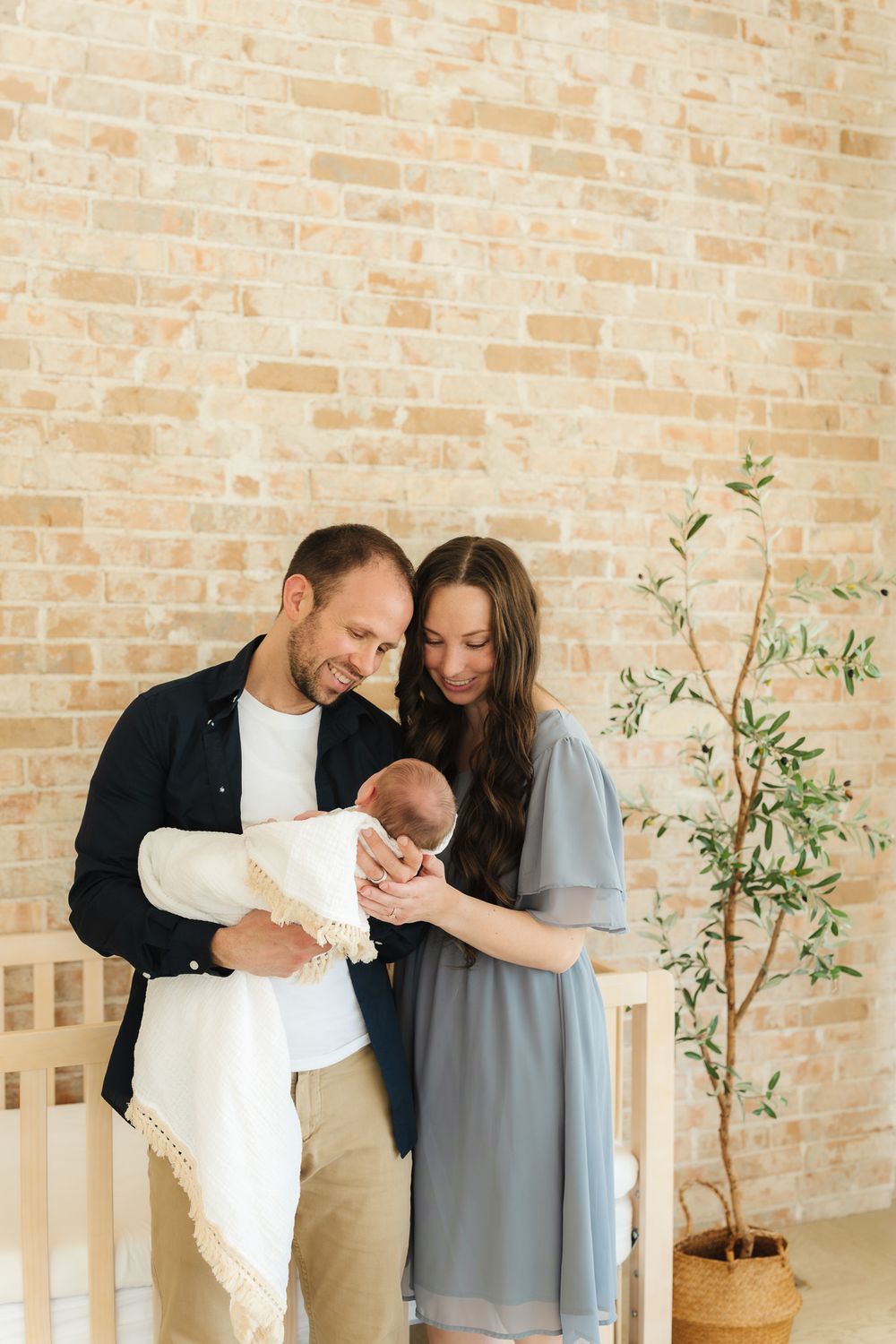 A couple stands lovingly with their newborn baby against a rustic brick wall with a small potted olive tree nearby.