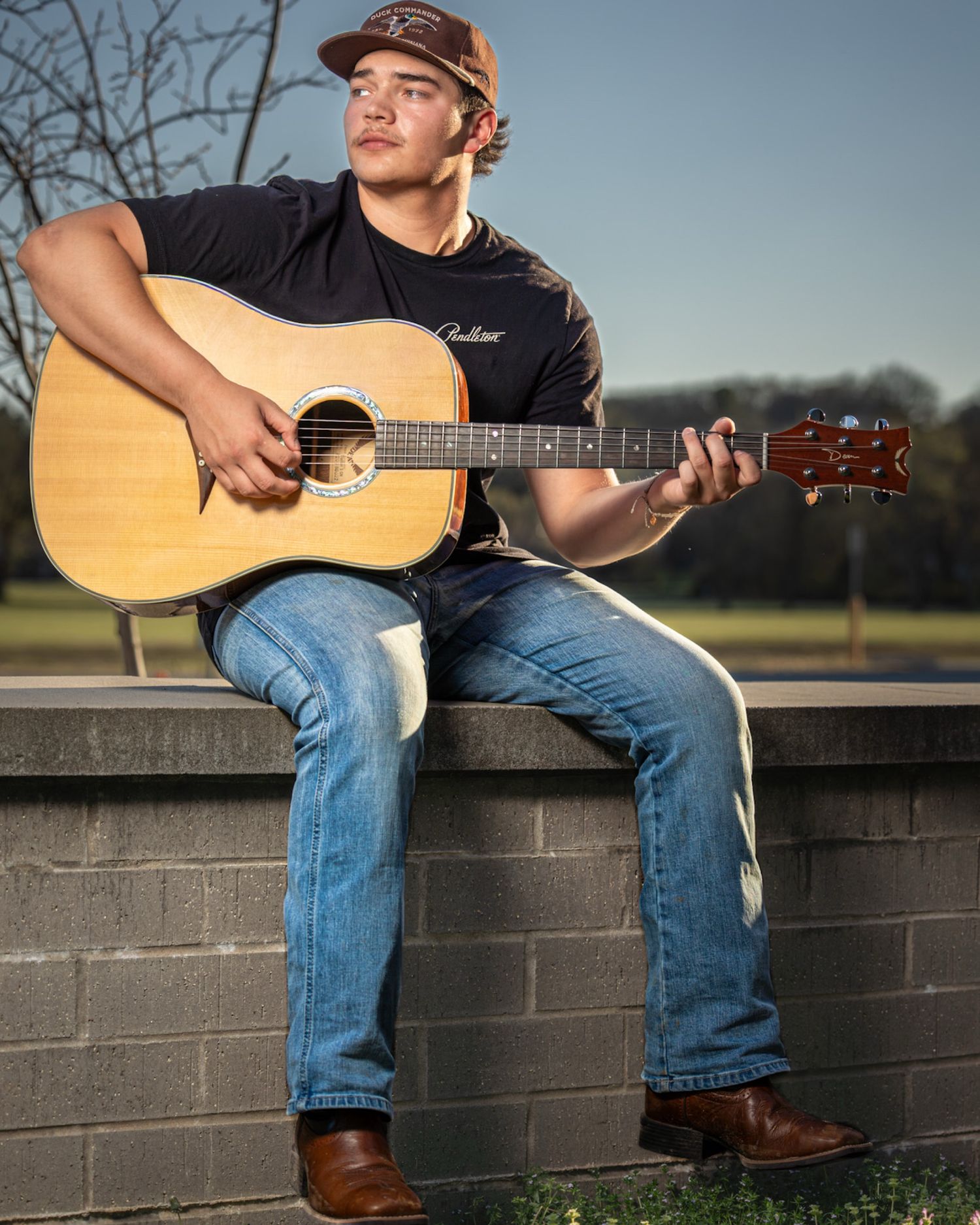 Person in baseball cap and jeans sits on brick wall playing acoustic guitar in outdoor setting at sunset.