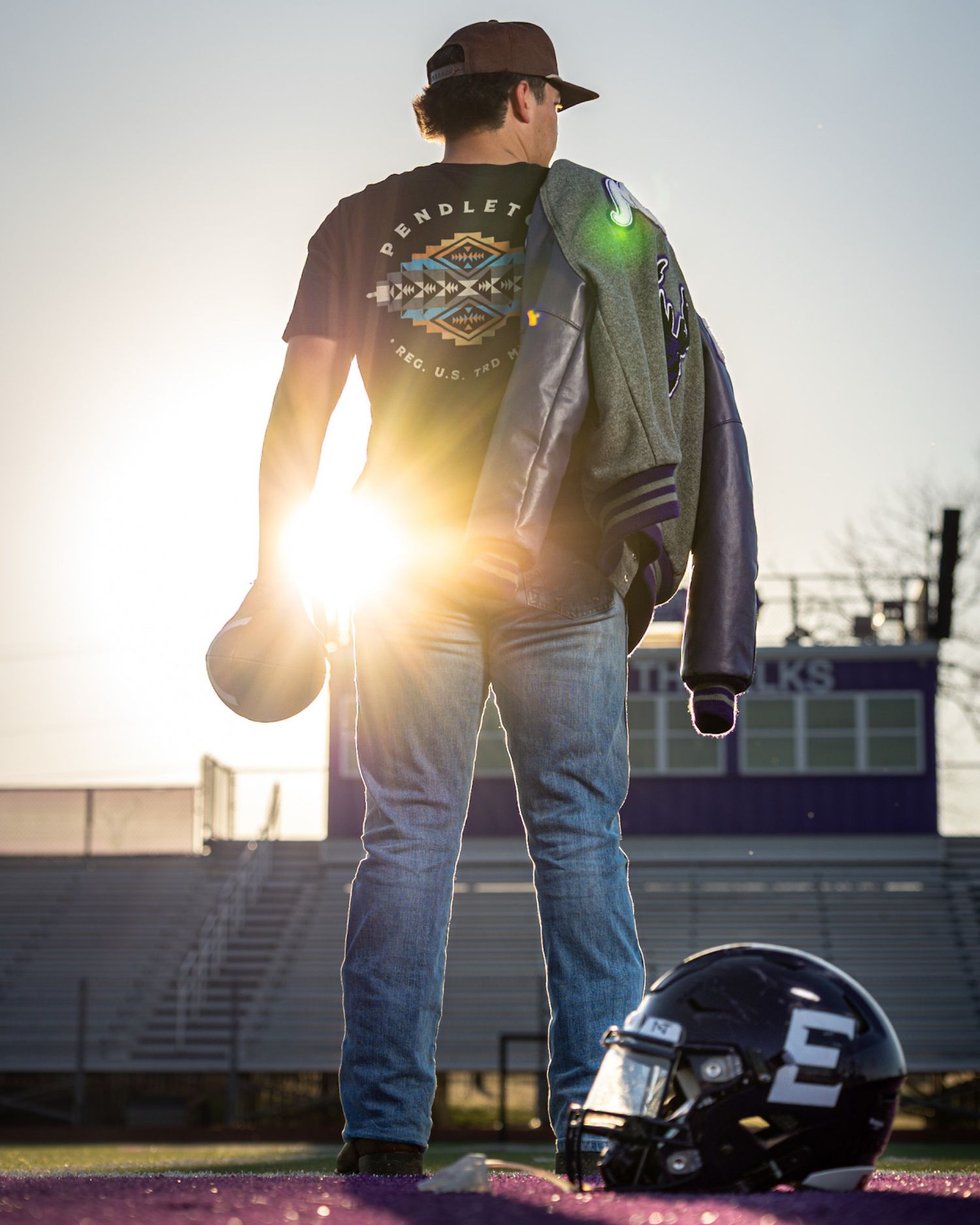 Person in jeans and dark jacket stands in silhouette against dramatic sunset at outdoor venue.